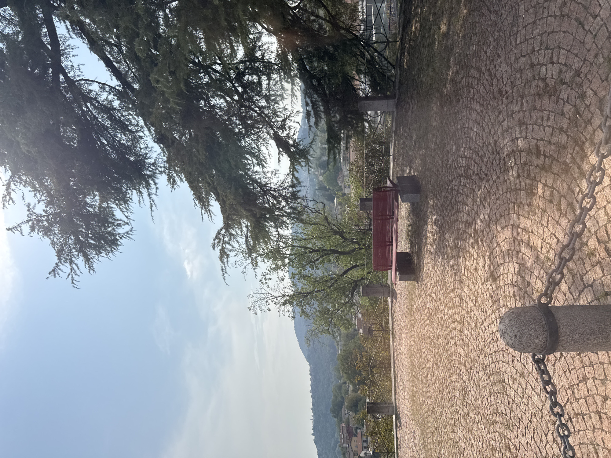 The image depicts an outdoor scene with a panoramic view of a town nestled in a valley, with hills in the background. In the foreground, a red bench is positioned on a cobblestone plaza, which is bordered by a low fence. A chain fence runs across the bottom left of the image, attached to a stone post. Large trees with dense foliage frame the upper right portion of the scene, casting shadows on the plaza. The sky is clear and blue with some light clouds. The location appears to be a viewpoint or park area, likely within or overlooking the town of Balerna, Switzerland, given the context. There are no people visible in the image.