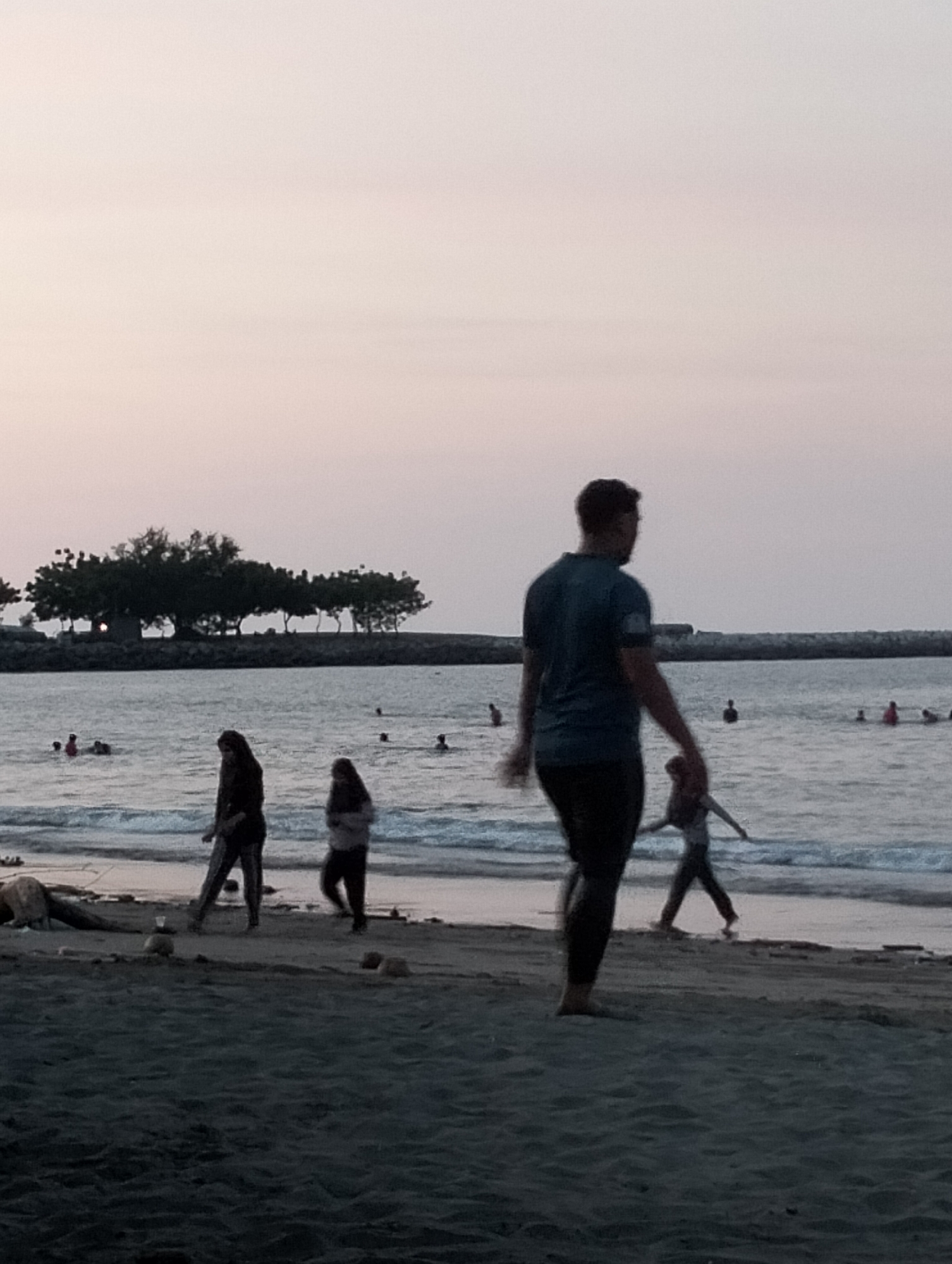 This image depicts a serene beach scene in Lhokseumawe, Indonesia, observed during dusk or late afternoon. The sky is a soft, diffused blend of pale pink, purple, and grey hues, indicating twilight.
In the foreground, a man, seen from behind and slightly out of focus, walks barefoot on the dark, wet sand towards the water's edge. He is wearing a dark t-shirt and pants, with his figure largely silhouetted. To his left, closer to the water, two women are strolling along the shoreline; one is dressed in dark clothing, while the other appears to be in lighter attire, possibly with a head covering. Further down the beach, a smaller figure, potentially a child, is visible near the water with arms outstretched.
The sea in the midground is dotted with several people swimming or wading, appearing as dark silhouettes against the water, with some showing hints of red clothing. In the background, a distant landmass features a cluster of dark, leafy trees, creating a natural horizon line, and a breakwater or rocky barrier is visible extending into the water. Scattered on the sandy beach near the water's edge are small, dark objects, possibly coconuts or stones, and a small white cup is also visible.
The overall atmosphere is peaceful and relaxed, capturing individuals enjoying a tranquil evening by the sea. No visible text is present in the image.