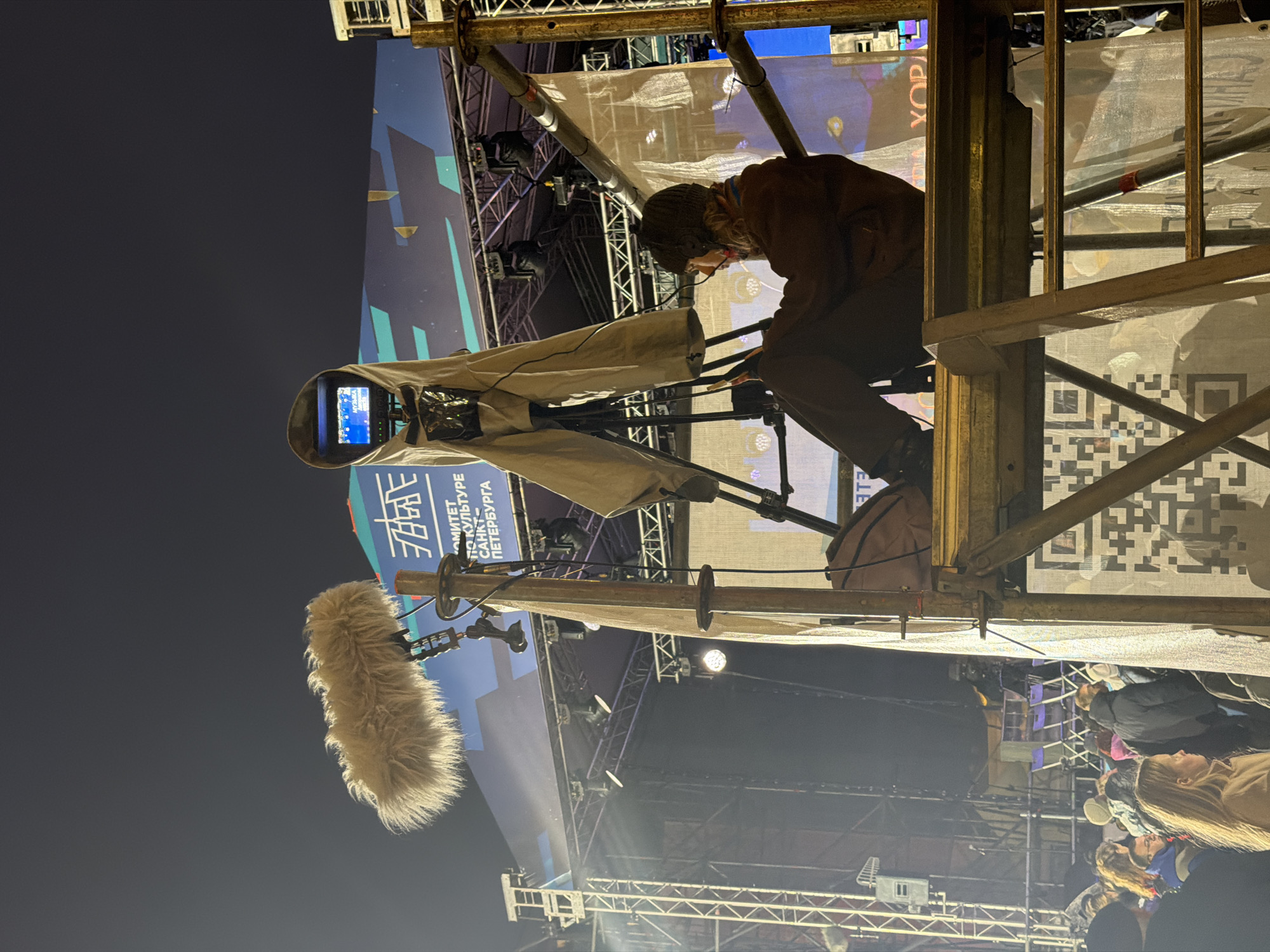 Here is a description of the image:
A camera operator is positioned high on scaffolding at an outdoor event in Saint Petersburg, Russia. The camera, covered in protective gear, is pointed towards a stage. A large furry microphone is visible. The operator is wearing dark clothing and appears to be monitoring the camera. A crowd of people is visible at the bottom of the frame. Banners with writing in Russian are in the background. The scaffolding structure shows the presence of a QR code.
