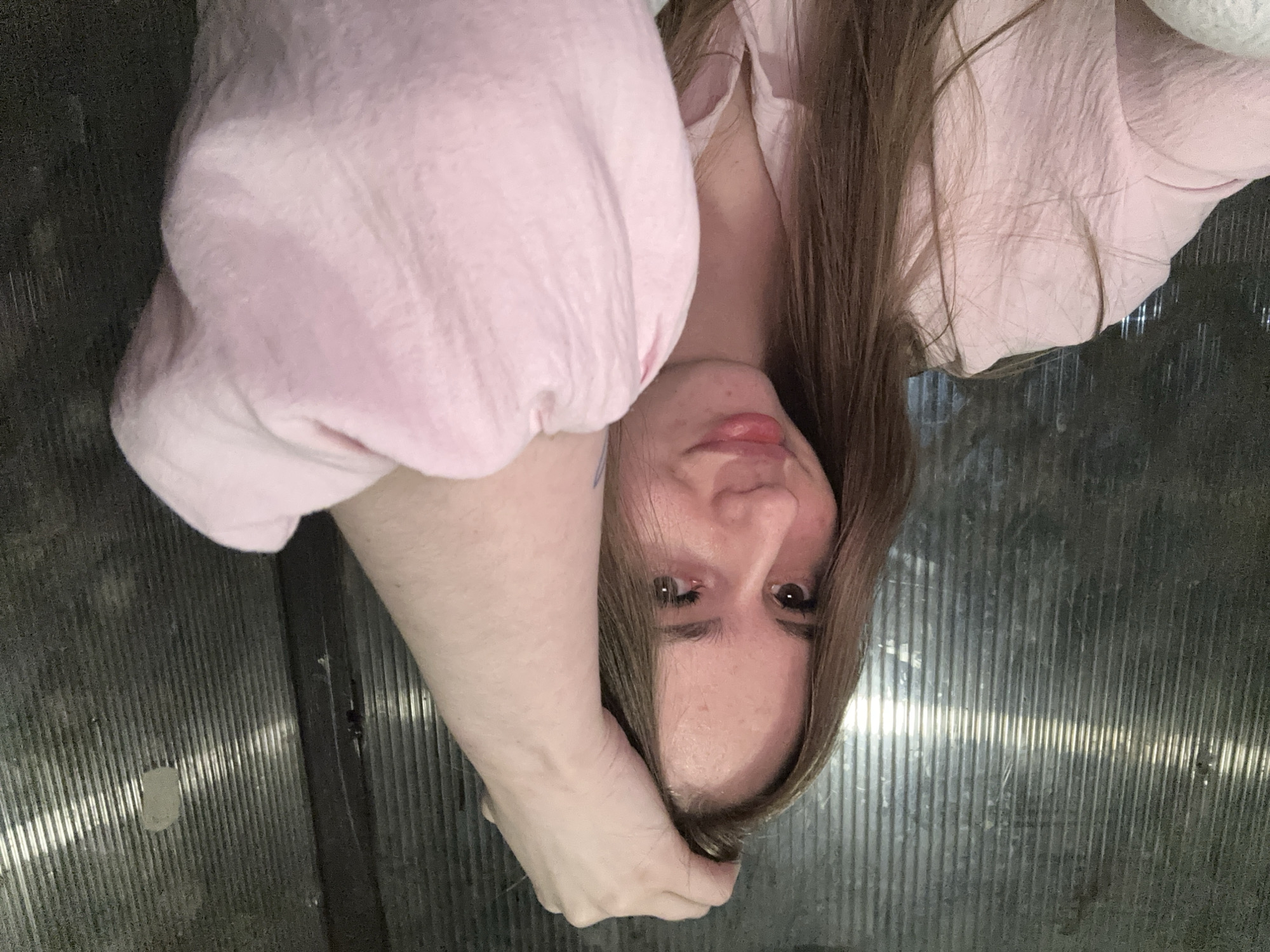 A close-up, horizontally oriented shot depicts a female subject lying down and looking directly at the camera. Her head is positioned towards the bottom of the frame, while her body extends upwards. She has long, straight brown hair that partially covers her forehead and right ear. Her dark eyes are open, and her lips are slightly parted.
She is wearing a light pink top with long, somewhat voluminous sleeves, appearing to be made of a soft, possibly satin or silk-like fabric. Her right arm is bent, with her hand supporting her head, and her left arm extends towards the upper left of the frame.
The background consists of a dark, corrugated or ribbed metallic surface, possibly a floor or wall. A bright horizontal line of light reflects across the middle of this surface, indicating an indoor setting with overhead illumination. The overall scene is simple, focusing on the individual's face and upper body in a relaxed pose against an industrial-like backdrop. No visible text is present.