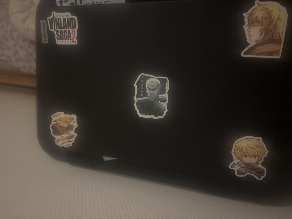 A black laptop is adorned with stickers. One sticker displays the "Vinland Saga 2" logo, while four others showcase anime characters. A profile view of a blonde character, a black and white depiction of an older man with a stern expression, and two different images of a younger blonde character in action poses are all present. A Lenovo logo is also visible on the laptop.