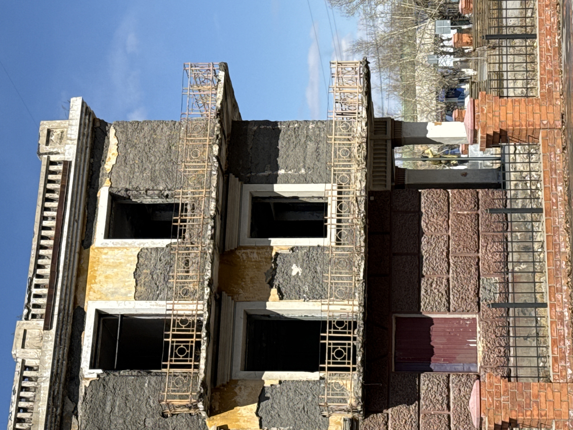 The image depicts a partially ruined building in Borzya, Russia. The building is a multi-story structure with visible signs of damage and disrepair. Sections of the facade are crumbling, exposing underlying materials. Window frames are present but lack glass, leaving dark, empty openings. Decorative metal grilles are affixed to the exterior of the building. The building appears to be a mix of concrete and brick. The surrounding environment includes trees and other buildings, indicating an urban or suburban setting. The sky is visible and is partly cloudy.