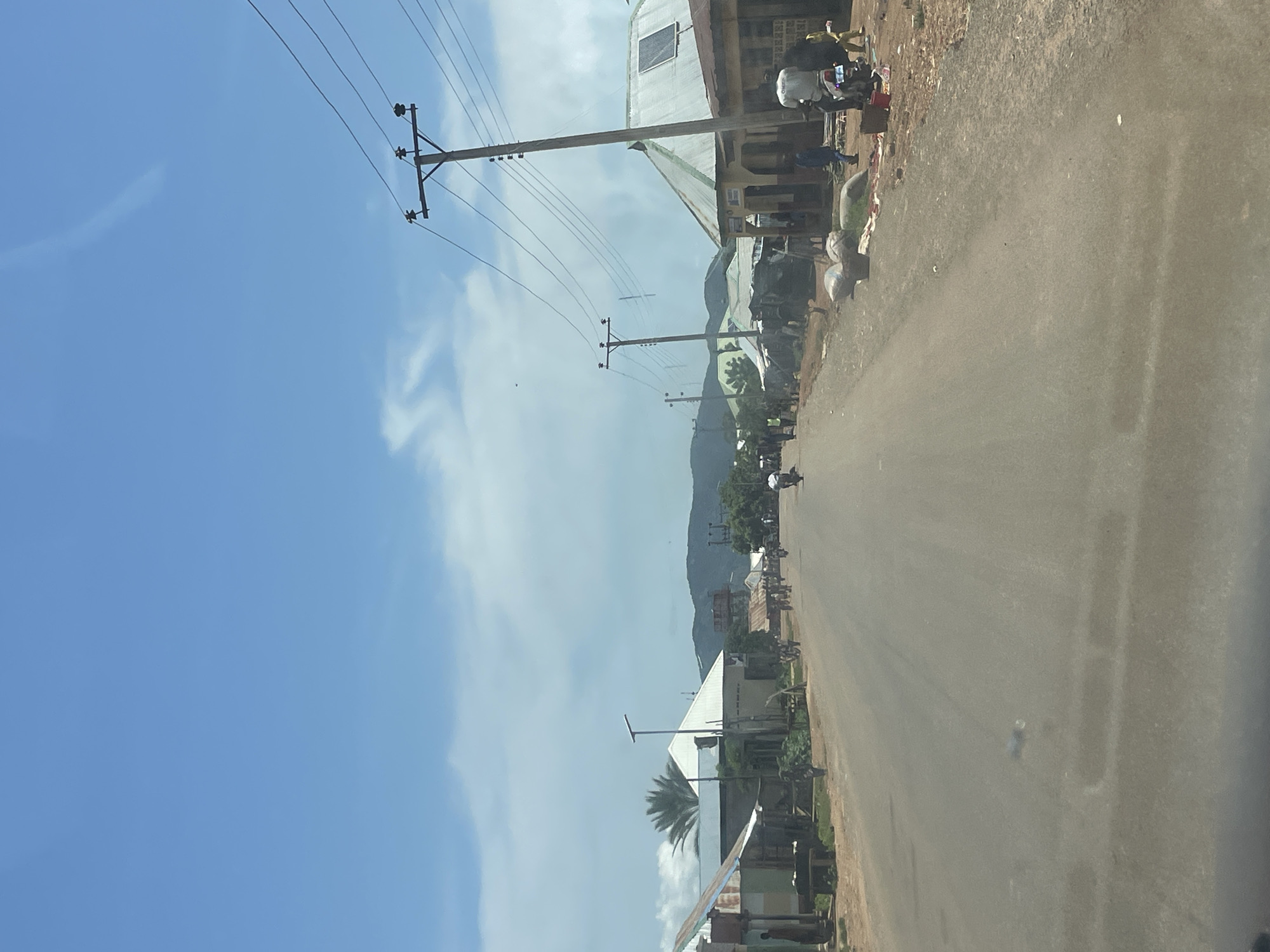 The image depicts a dusty road in a town, likely within the city of Zalau, Nigeria. The road is paved and appears to be a central thoroughfare. On either side of the road, there are various structures, including buildings with corrugated metal roofs, some appearing to be shops or homes. Power lines are prominently visible, supported by wooden utility poles.
Several people are present in the scene. Some are walking along the road or standing near the buildings. There are also individuals on motorcycles, suggesting a common mode of transportation in the area. Some of the buildings appear to have goods displayed outside, possibly indicating market activity or informal commerce. Bags of materials, possibly agricultural products or merchandise, are stacked along the roadside. In the distance, hills or mountains are visible, adding to the natural landscape surrounding the town. The sky is blue with scattered clouds, indicating a daytime scene. The overall impression is one of a busy, functional street in a Nigerian town, with elements of daily life, commerce, and infrastructure.