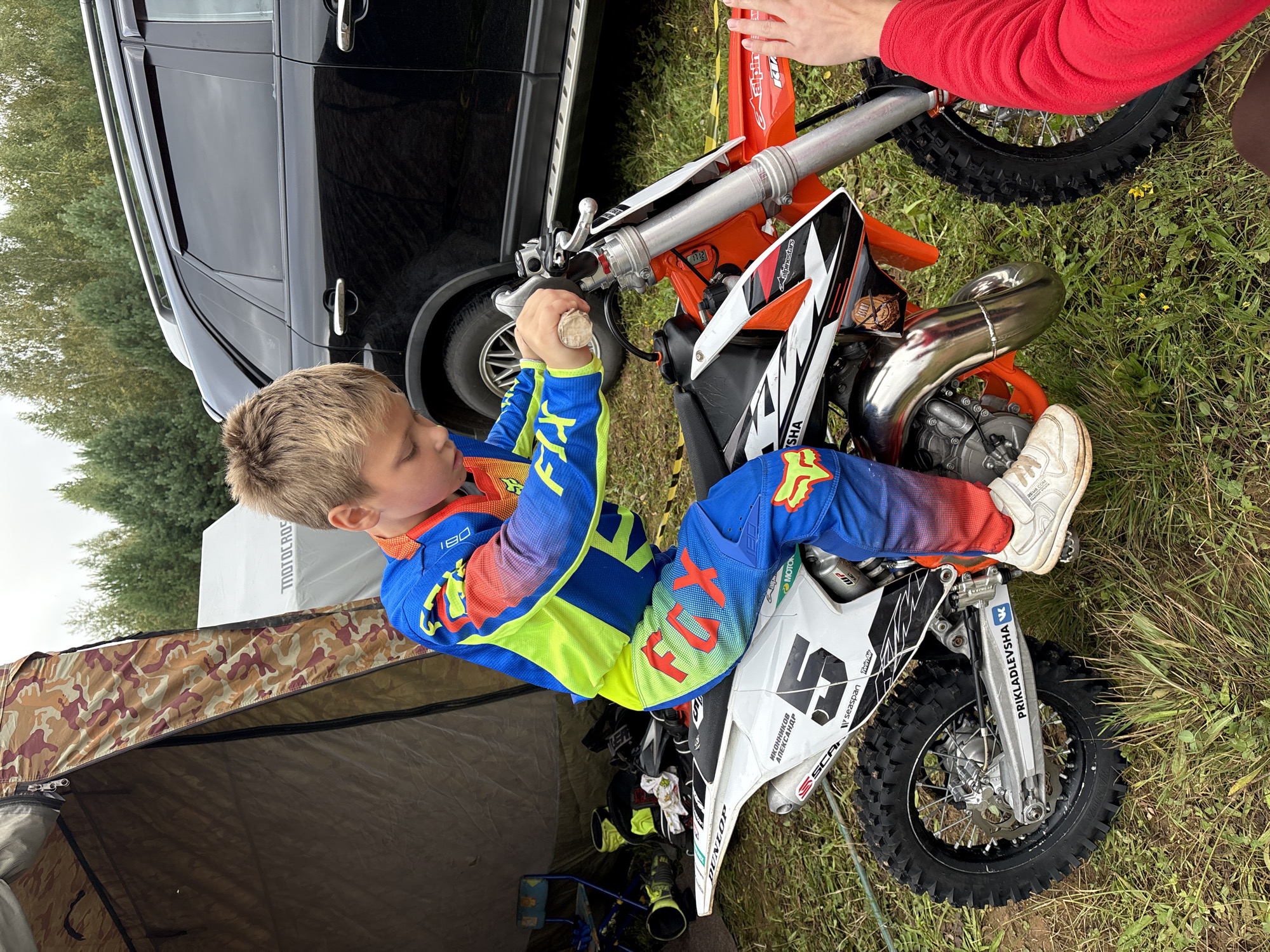 A young Caucasian boy wearing a blue and neon yellow motocross suit with orange accents is sitting on an orange and white dirt bike. He is looking down at the handlebars, which he is holding with both hands. The boy has blond hair and is wearing white sneakers. He is positioned in front of a camouflaged tent on the left side of the frame, with a black SUV in the background on the right. The scene appears to be outdoors, with grass and trees visible. Text on the dirt bike includes "FOX", "DUNLOP", "SCAP", "PRIKLADEVSK", and "SEASPAN". The image is taken from a slightly low angle. The overall impression is a casual outdoor activity, likely a motocross event or practice session. There is no specific indication that this location is within the city of Balak, Russia.