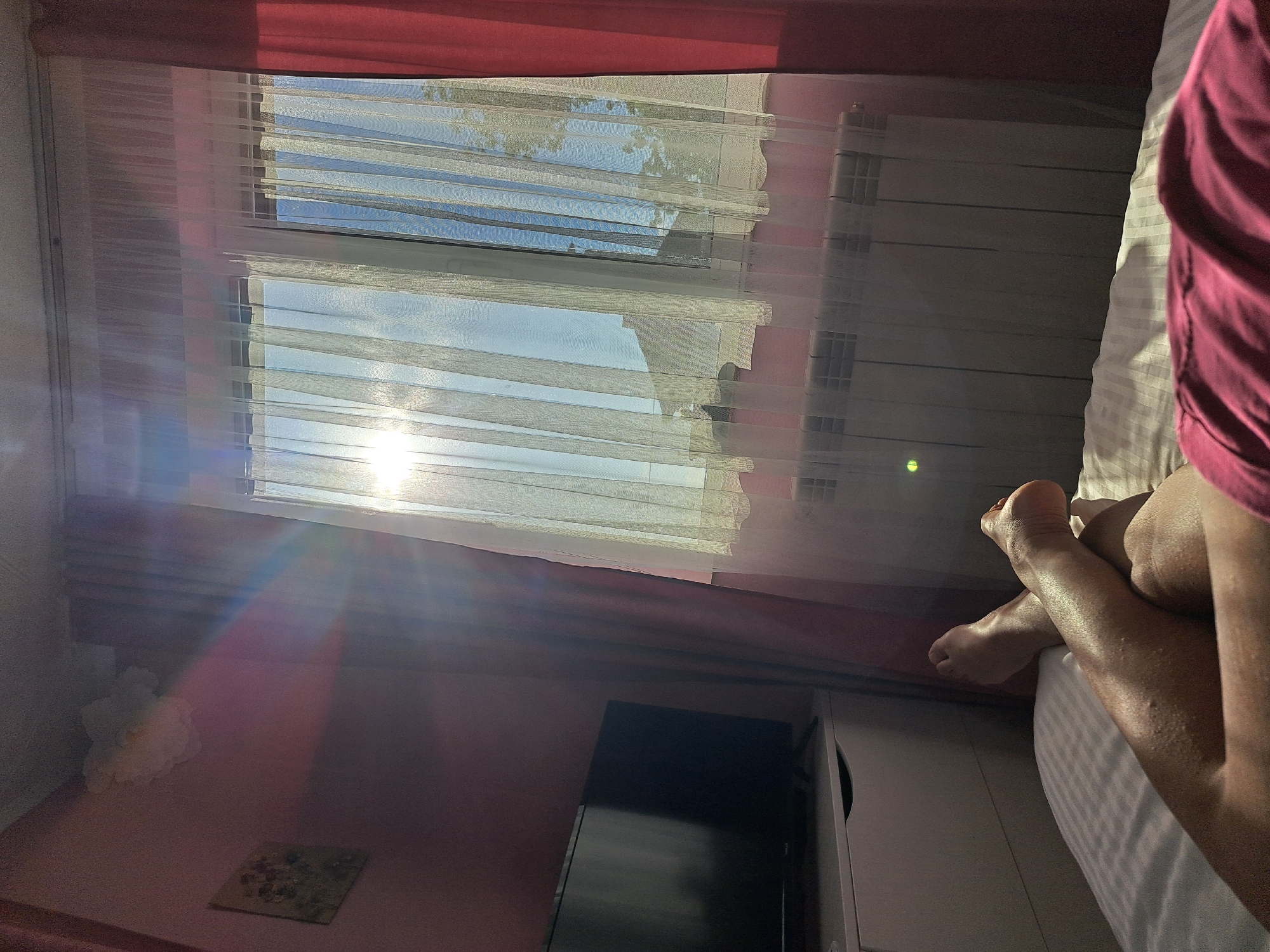 Here is a description of the image:
The image shows an interior scene with a view of a window covered by sheer curtains. Sunlight streams through the window, creating a bright glare and illuminating the room. The window reveals a view of the outside, possibly with a blue sky visible between the slats. A radiator is visible next to the window.
To the side, a person's legs and feet are visible on a bed, partially covered by a striped bedsheet and a pink garment. A television is also visible. The wall is painted in a shade of pink, and there is a piece of decor on it. The location is unknown, and the scene is a domestic interior.