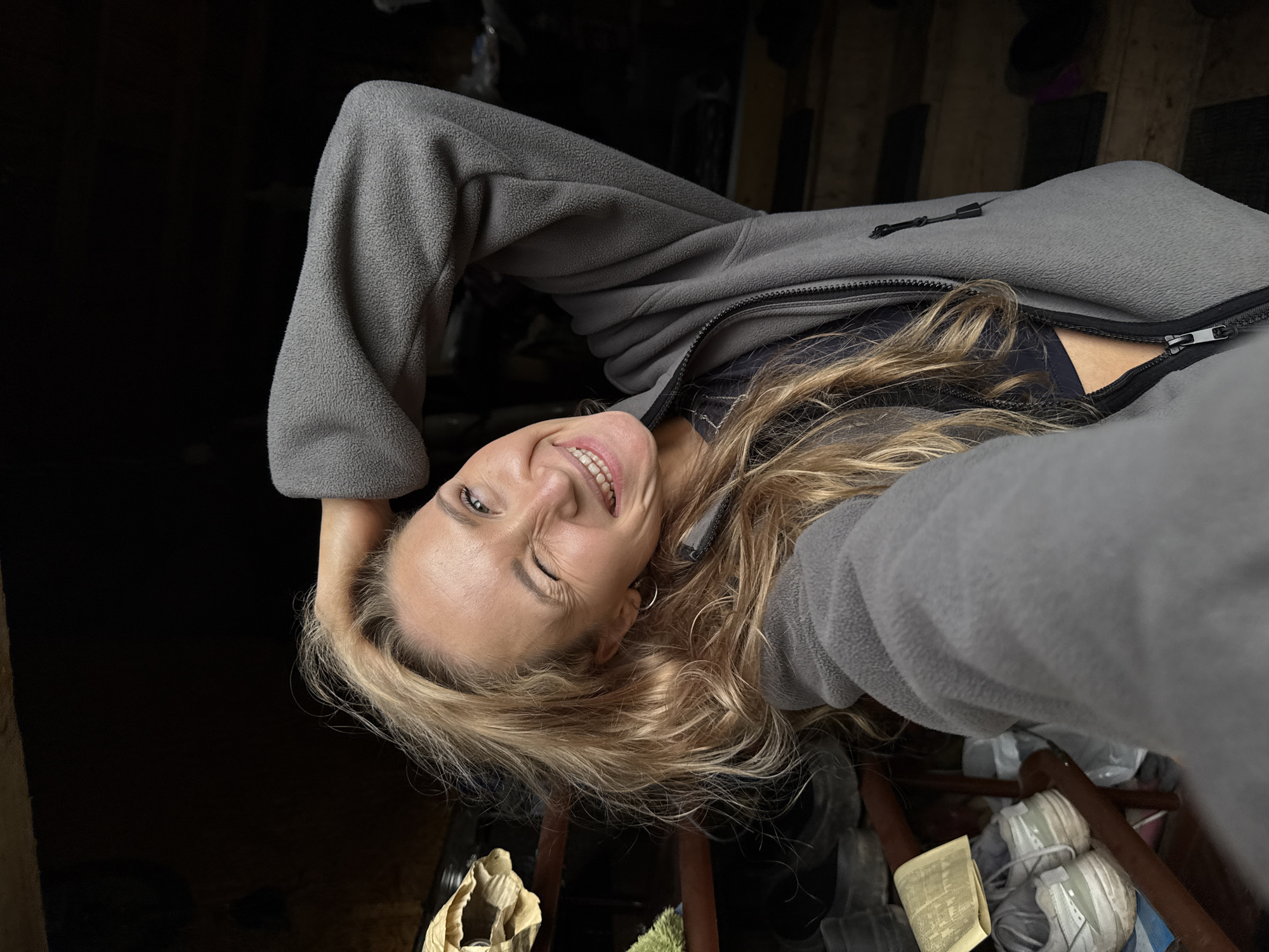 A young Caucasian woman with blonde, wavy hair, wearing a gray fleece jacket and a dark sports bra, is taking a selfie. She is indoors, in what appears to be a dimly lit storage area. Her left hand is raised, touching her hair, and she is winking at the camera with a wide smile. The background is dark and cluttered, with shelves holding various items, including what appear to be shoes and bags. The setting is not identifiable as being in the city of Nizhnyaya Chermoda, Russia.