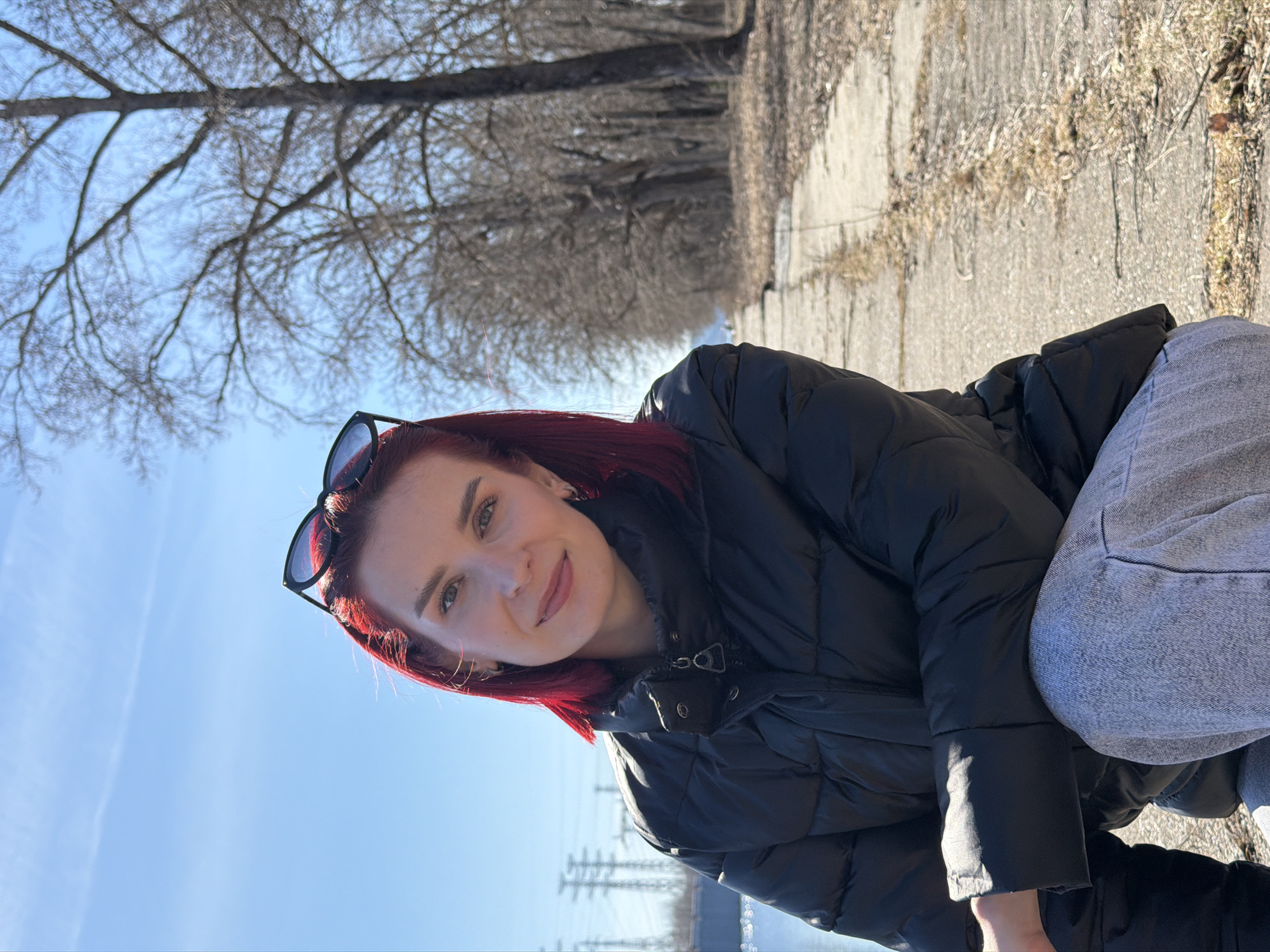 A woman with red hair and sunglasses is crouching outdoors on a paved surface. She is wearing a black puffer jacket and light gray jeans. Behind her, trees line the paved area, leading into the distance. The sky is blue. In the far background, power lines are visible. The location is the city of Chaikovsky, Russia.
