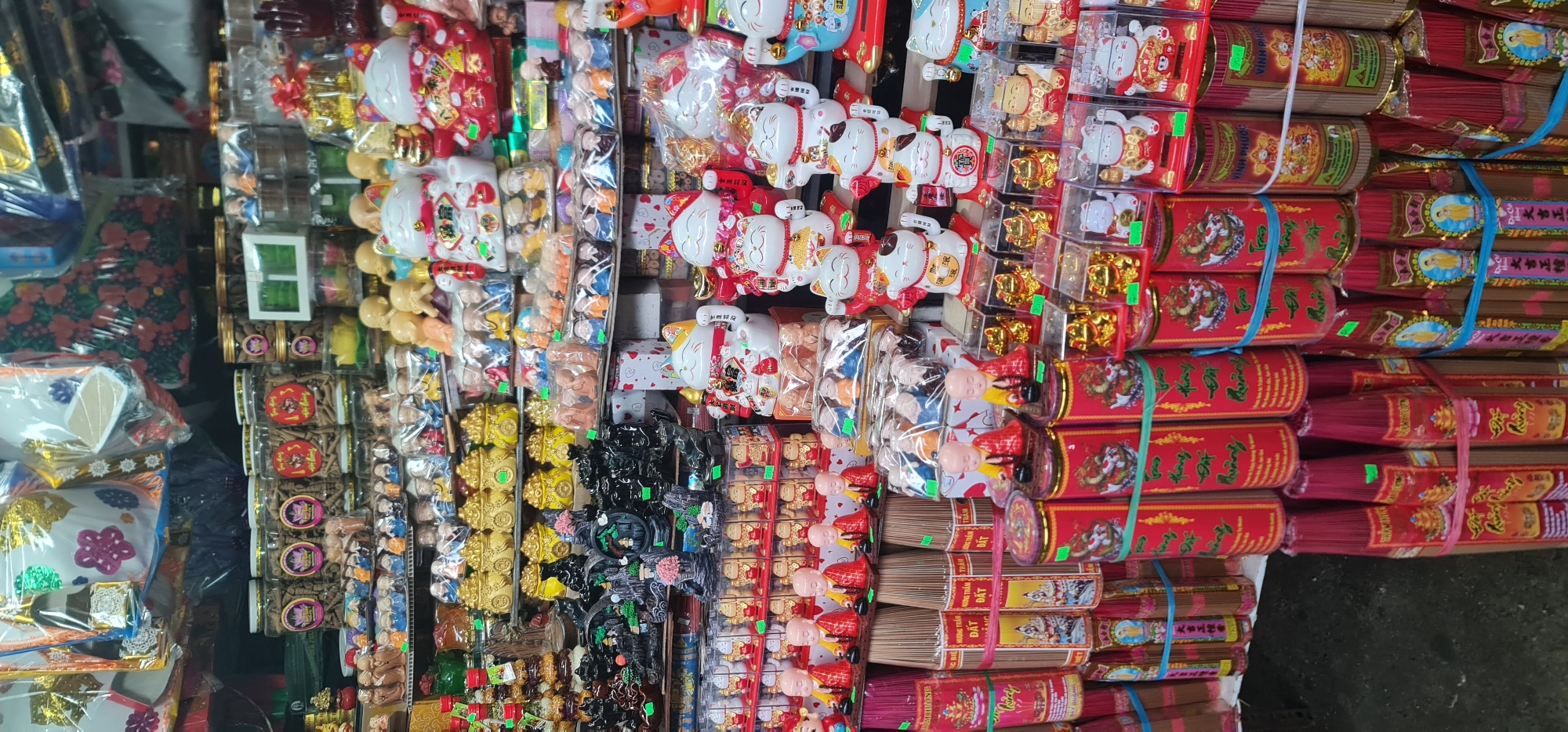 This photograph shows a vendor's stall in a market in Đà Nẵng, Vietnam. The shot is oriented vertically, showcasing a variety of products intended for religious or ceremonial use.
On the right side, stacks of red incense sticks are held together with blue and green rubber bands. The packaging has Vietnamese and Chinese characters, implying the origins of the product, along with a Buddha image. To the left of the incense sticks, there are plastic-wrapped figurines, including cats, gold objects, and small monks, commonly associated with prosperity, luck, and protection in Asian cultures. Further to the left, more packaged items are visible, including boxes containing incense and decorative pieces, such as flowers and gift items wrapped in plastic.