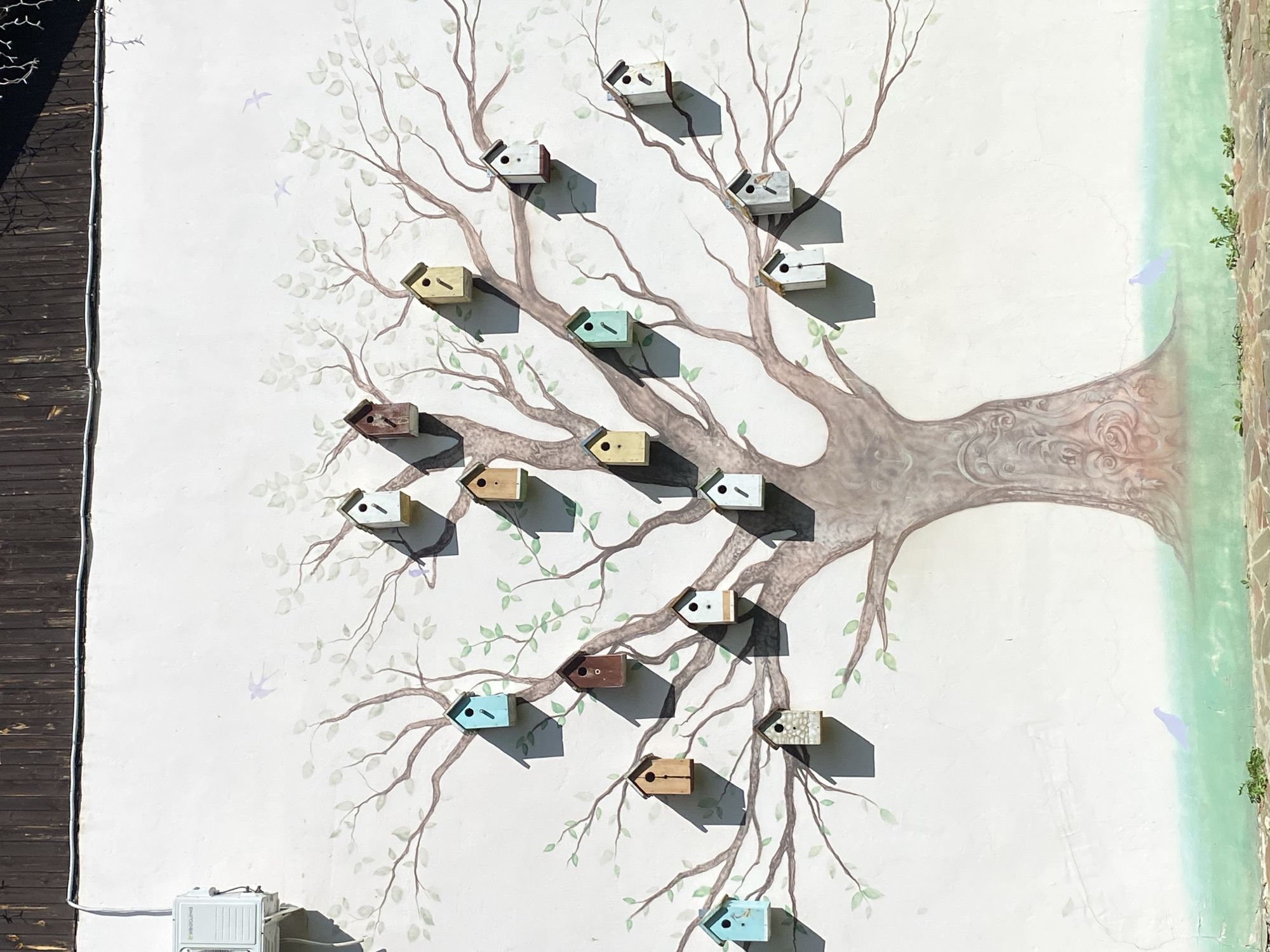 The image depicts a mural painted on a white wall, featuring a large tree with numerous birdhouses attached to its branches. The tree is rendered in shades of brown and gray, with delicate green leaves scattered throughout. The birdhouses are small and varied in color and design, some appearing to be made of wood, others of stone. The mural also includes faint outlines of birds in flight and at the base of the tree, painted in a light purple hue, against a background of green grass and a stone border. An air conditioning unit is visible on the upper left side of the wall, mounted above the mural. The scene appears to be outdoors, under a bright sky, with shadows cast by the birdhouses. The mural is located in the city of Chulis, Russia.