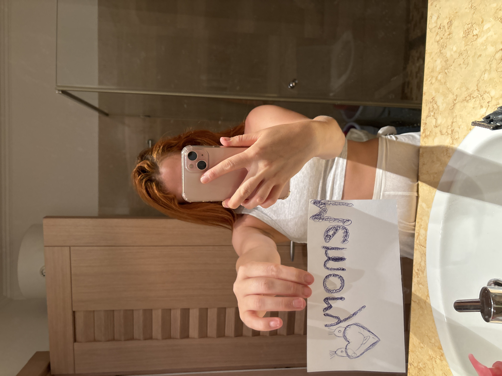 A person, appearing female with reddish-orange hair, is depicted taking a mirror selfie in a bathroom located in Budva, Montenegro. The individual is wearing a light grey crop top and light-colored bottoms. The subject's right hand holds a light-colored smartphone (an iPhone with a triple camera array) directed towards the mirror, while the left hand holds a white rectangular sheet of paper. The paper displays the mirrored word "Mom" written in blue pen with a bubbly or dotted texture, accompanied by a blue pen drawing of a heart with a face and two small bird-like figures positioned above it. The bathroom environment, reflected in the mirror, includes light-colored wooden cabinetry featuring horizontal slat details, a light beige marbled countertop, a white sink with a chrome faucet, and a partially visible pink soap dispenser. A shower enclosure with dark-tiled walls and a glass panel is reflected on the right side of the image, and a white circular fixture is visible on the wall above the cabinetry.
