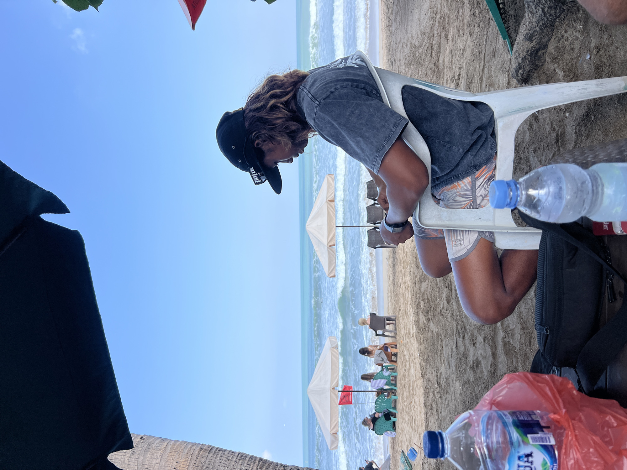 A person is sitting on a white plastic chair at Kuta Beach in Kuta, Indonesia. The person is wearing a dark t-shirt, patterned shorts, and a black baseball cap. They are looking down at a smartphone in their hands. In the foreground, two plastic water bottles and a red plastic bag are on a dark surface, possibly a table. The beach is sandy, and there are several beach umbrellas and chairs in the middle ground. The ocean with waves is visible in the background under a clear blue sky.
