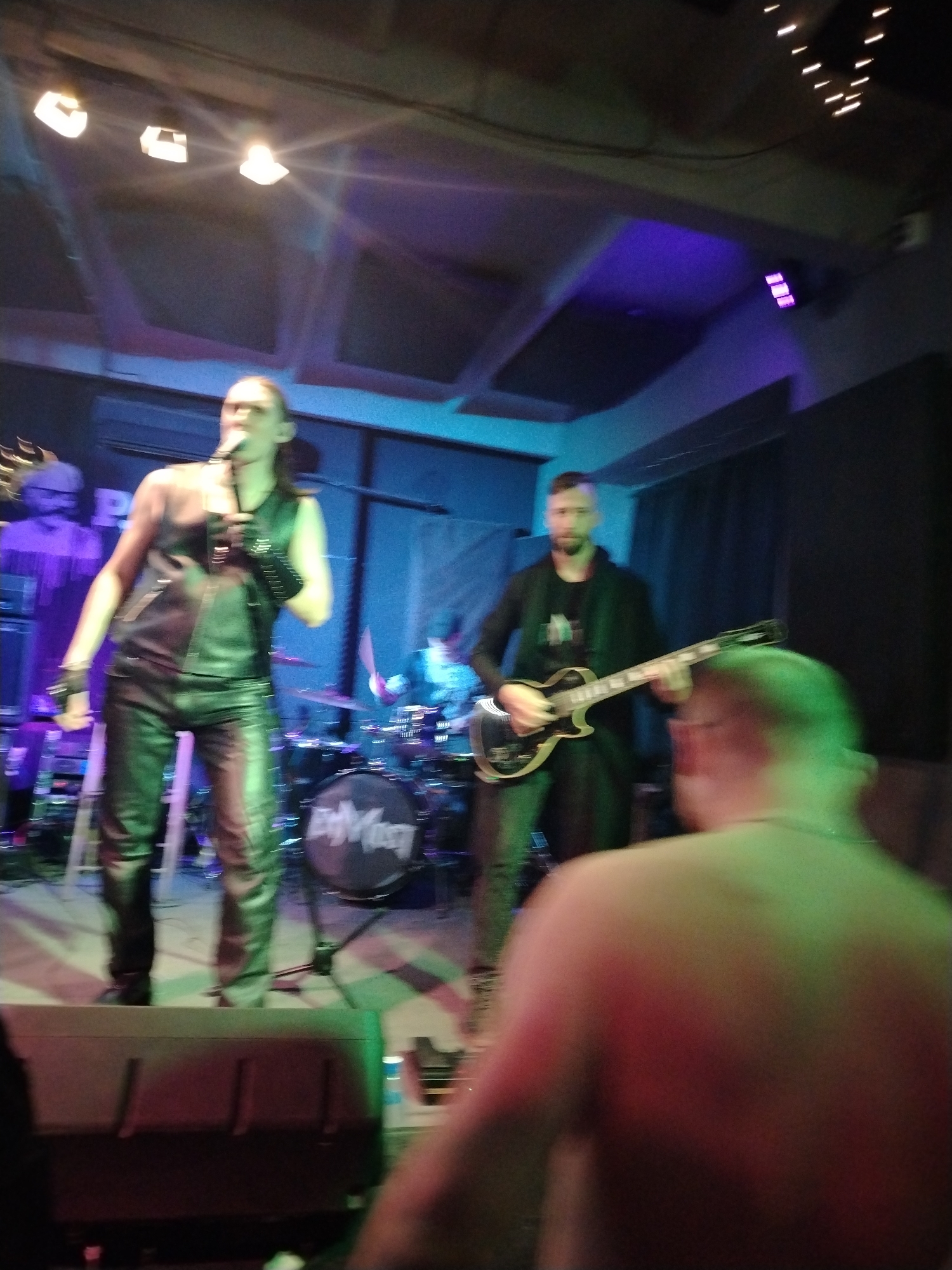 A band is performing on a stage in what appears to be a music venue. The lead singer is on the left side of the stage, wearing a black leather vest, black leather pants, and black leather gloves. He is holding a microphone and singing. A guitarist is standing to the right of the singer, wearing a black blazer and jeans and playing a black electric guitar. In the background, behind the singer and guitarist, a drummer is playing. The drum set has the band's name on the bass drum. The stage is lit with blue and purple lights. In the foreground, a blurred person, shirtless, is visible. It's difficult to determine the specific location in Chaikovsky, Russia, without additional details.