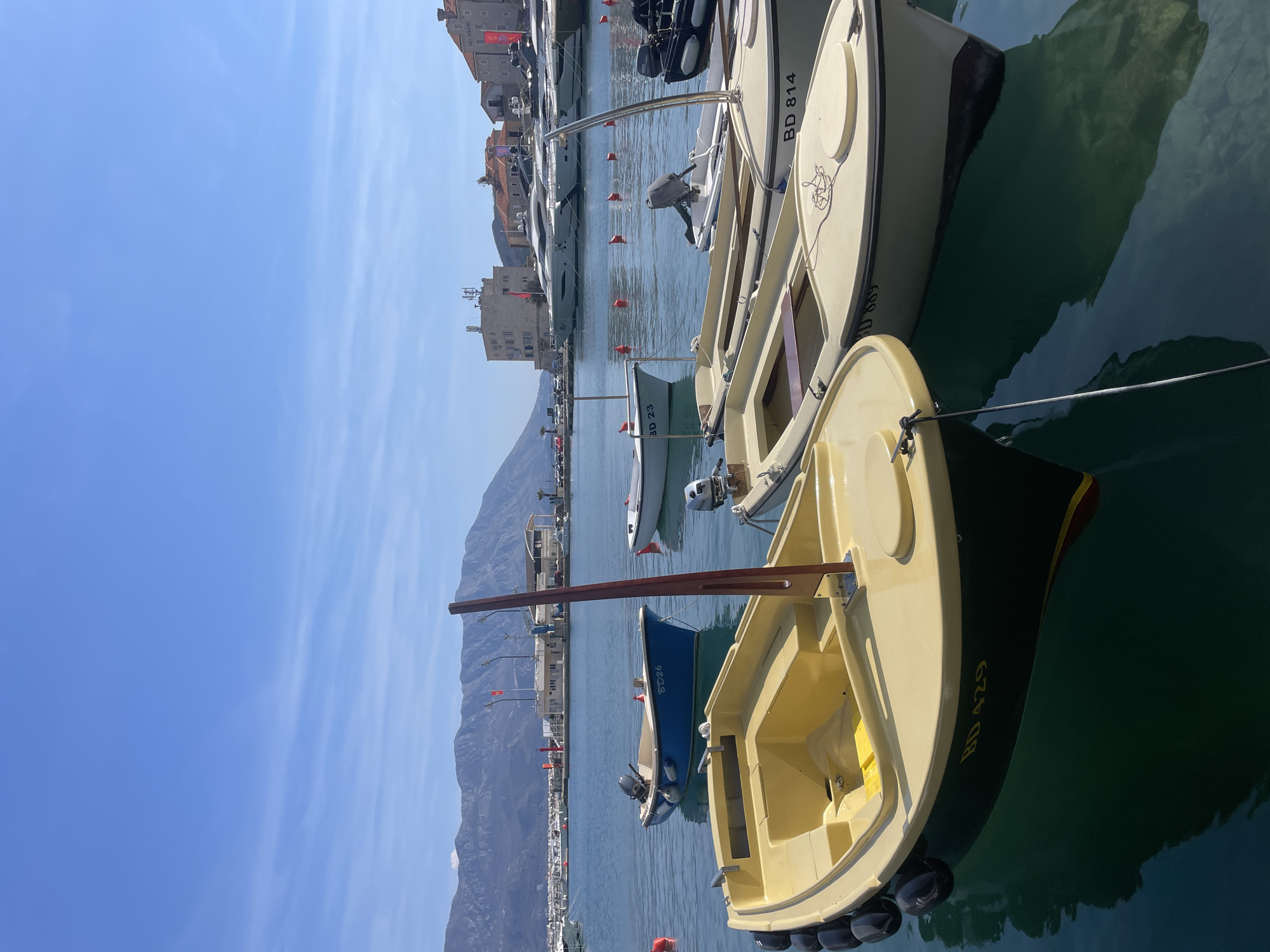 The image depicts a marina in Budva, Montenegro, under a clear, bright blue sky with sparse, wispy cirrus clouds. The scene is dominated by numerous small boats moored in calm, greenish-blue water that reflects the sky and vessels.
In the immediate foreground, a light yellow boat with a dark green hull is visible on the left, displaying the registration "BD 429" in yellow text. It features black fenders along its port side and a brown wooden mast-like structure protruding from its interior. To its right, partially obscured, is another light-colored boat with "BD 814" visible on its bow, and "BD 869" partially visible below it on the hull, suggesting a boat directly behind it. This boat has a coiled rope on its foredeck and an outboard motor mount at the stern.
In the midground, a blue boat with a white interior, bearing the registration "BD 26," is moored with an outboard motor attached. Further back and to its right, another light-colored boat marked "BD 23" with an outboard motor is visible. Several red cylindrical buoys are scattered across the water. To the far right, a black inflatable boat with an outboard motor is partially visible.
The background features a range of elements. On the left and center, a modern waterfront building, possibly a marina office or restaurant, is visible, with multiple large, white luxury yachts docked beyond the smaller boats. Red flags are present on poles along the waterfront. Further back, vehicles are parked along a road. To the right, historic stone buildings with red-tiled roofs, characteristic of Budva's Old Town, are visible along the shoreline. A range of distant mountains under the clear sky forms the horizon. No individuals or specific human actions are discernible within the frame.