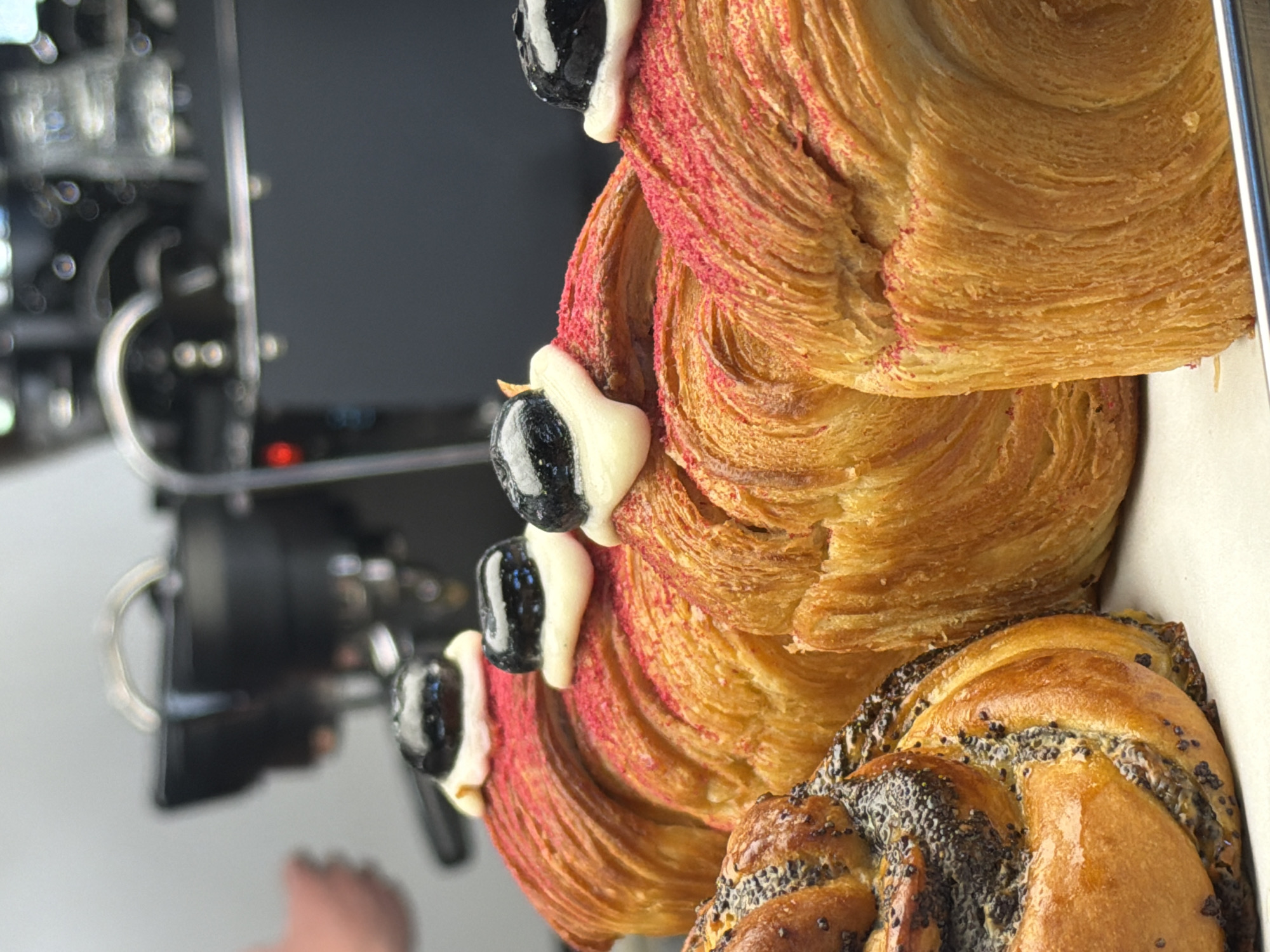 The image shows a close-up of several pastries in a container, likely in a bakery setting. The topmost pastry is a croissant-like pastry with a swirled, layered texture. It is topped with a red-colored dusting or coating and adorned with black cherries held in place by white cream. Beneath it is another similar pastry, and at the bottom is a braided pastry covered in poppy seeds.
A blurry, dark-colored machine is visible in the background, suggesting the presence of other bakery equipment. A hand or partial foot is also visible in the lower-left corner of the image.
There is nothing in the image to suggest the location is in the city of Izhevsk, Russia.