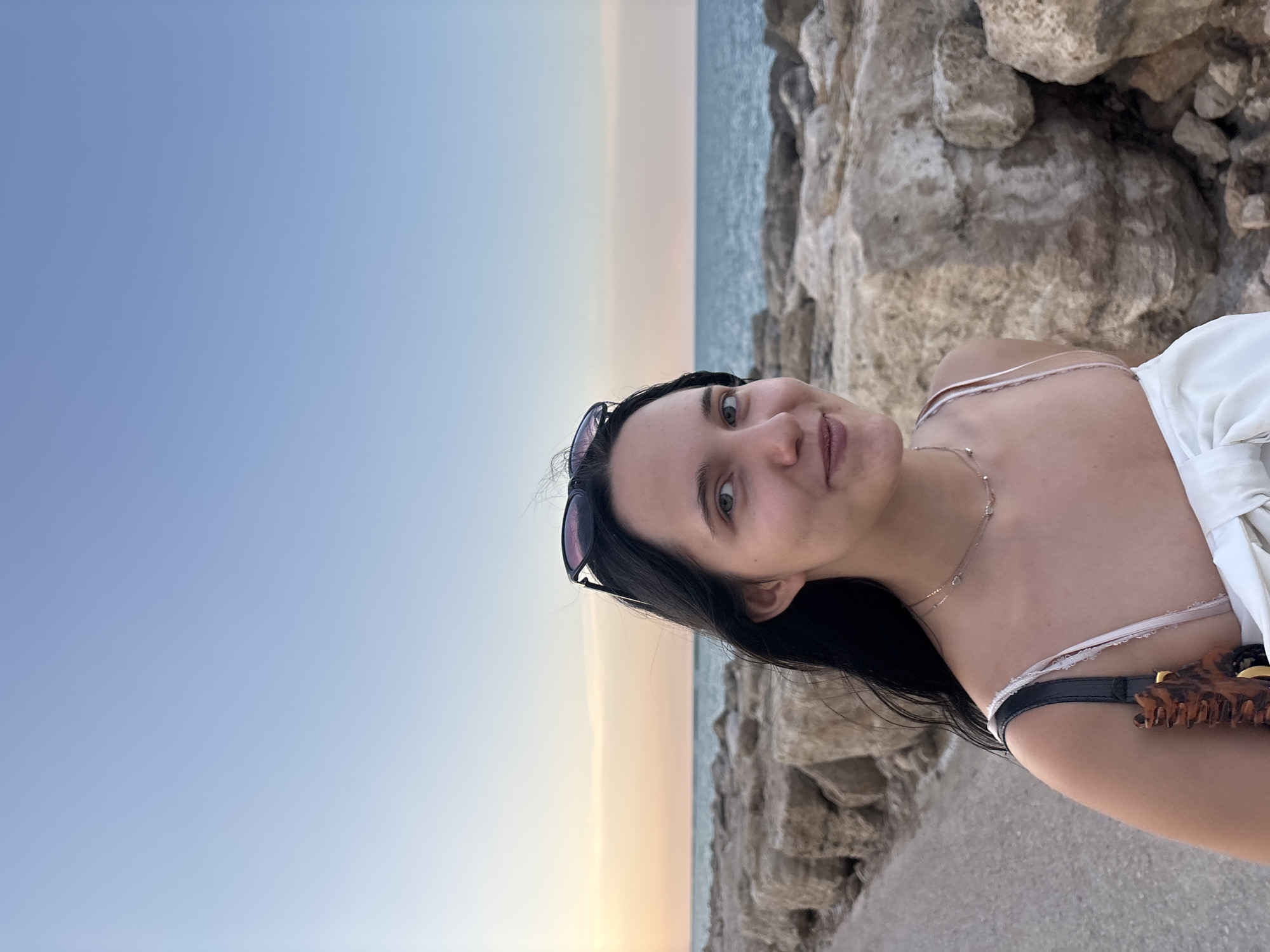 The image shows a young woman standing on a rocky embankment near the sea, possibly in Grottammare, Italy, during sunset. The sky is a gradient of light blue to pale orange, indicating the sun is low on the horizon. The woman has dark hair, sunglasses perched on her head, and is wearing a white dress with thin straps. She is looking towards the camera with a slight smile. The foreground is dominated by large, rough-textured rocks, and a paved pathway is visible to her left. The sea stretches out behind her, with a clear horizon line. The overall mood is serene and captures a moment of leisure by the coast.