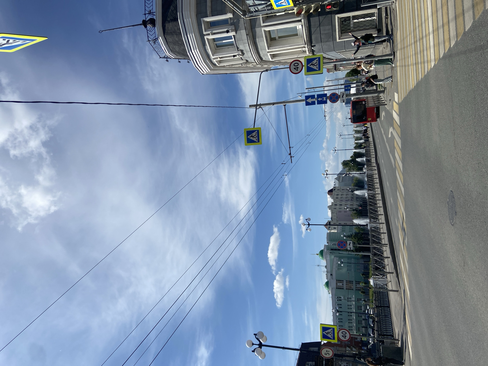 This image captures a street scene in Kazan, Russia, on a clear, sunny day with a blue sky and scattered white clouds. The perspective is from the road, looking forward along a multi-lane asphalt street with a marked pedestrian crossing in the foreground.
To the right, a multi-story building with a distinctive rounded corner features a light grey facade, multiple windows, and a dark, ornate roof with a decorative railing. A traffic light showing red is mounted on its corner. Three individuals are on the sidewalk near this building: one person in light clothing stands closer to the building, another in dark clothing has their arms raised, and a third in green clothing stands between them, all facing generally towards the street. Overhead utility wires descend diagonally towards a tall street pole in front of this building, displaying two "pedestrian crossing" signs (blue square with white triangle and black figure), a "40" km/h speed limit sign, and two "straight ahead" directional arrow signs. Another "pedestrian crossing" sign is partially visible at the top right of the frame.
Along the left side of the street, a metal railing separates the road from a sidewalk. Further along this sidewalk, fountains are visible spraying water. Buildings with varying architectural styles and colors, including a prominent light green building with green dome-like structures on its roof, line the far side of the street. A person in dark attire stands on the sidewalk near the railing on the left, next to a dark utility box. On the left, a street sign indicates a "5T" weight limit, accompanied by another "pedestrian crossing" sign and a "40" km/h speed limit sign. Multiple streetlights with spherical lamps are positioned along both sides of the street. Overhead wires stretch across the entire upper portion of the sky. In the mid-ground, a red public transit bus is visible heading away from the viewer on the right side of the road. A manhole cover is present in the foreground of the road.