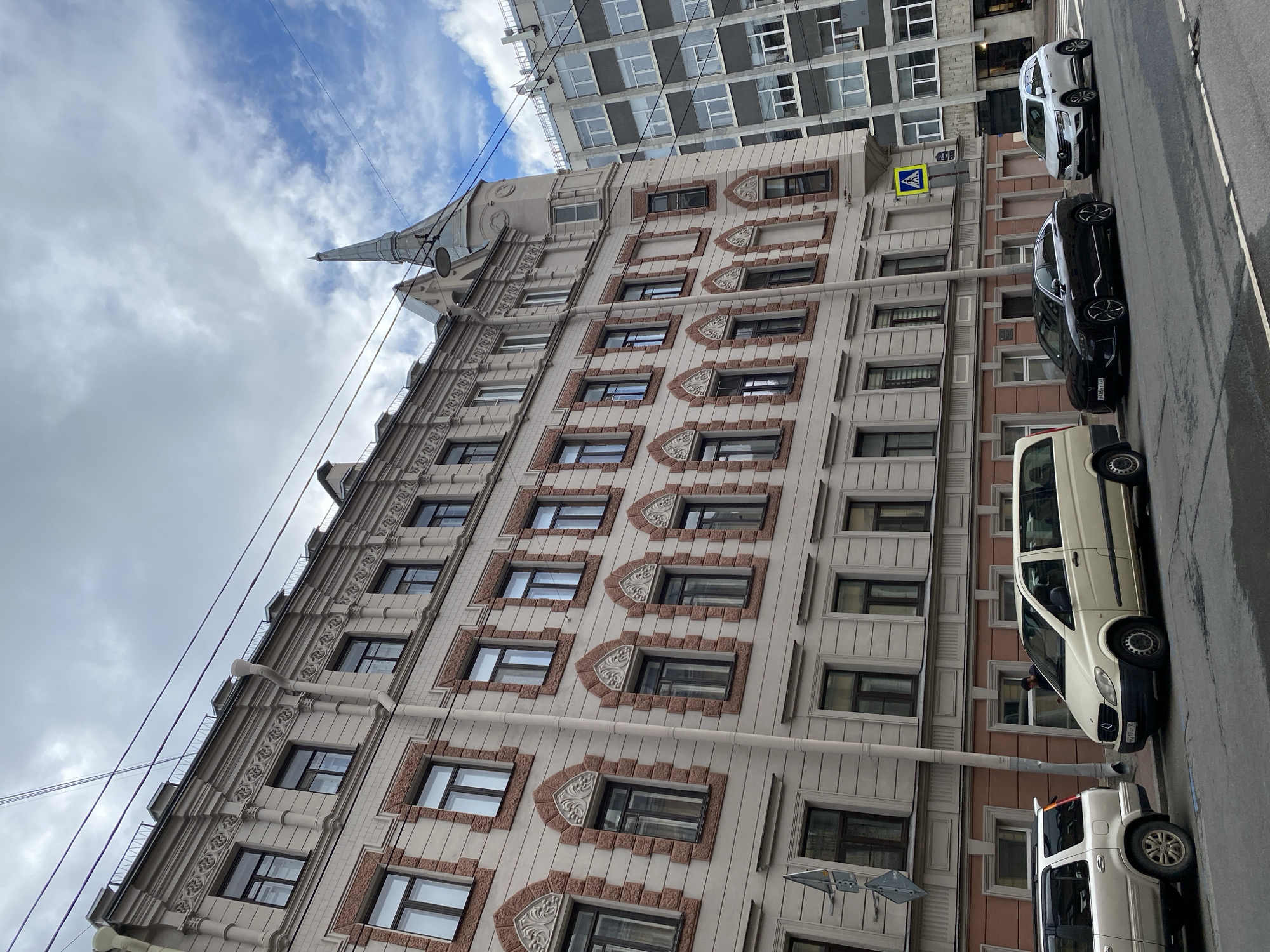 The image shows a street scene in Saint Petersburg, Russia. A multi-story building with light pink and grey facade, decorative brickwork around the windows, and an ornate cornice dominates the left side of the frame. A pointed spire-like structure is visible on the corner of this building. To the right, a modern grey building with many windows is visible. Several cars are parked on the street: a silver SUV on the left, a cream-colored minivan in the middle, a black sedan, and a silver SUV on the right. A pedestrian signal with a yellow diamond shape and a white pedestrian symbol is mounted on a pole near the sidewalk. The sky is partly cloudy with patches of blue. Overhead power lines are visible crossing the sky.