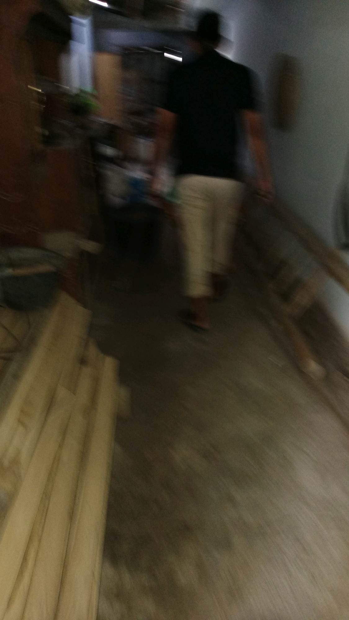 The image is a blurry, low-resolution photograph taken indoors, possibly in a workshop or storage area. A person, silhouetted and out of focus, is walking away from the camera. They are wearing a dark t-shirt and light-colored pants, and a dark hat. To the left of the person, there are stacks of wooden planks, a dark bin, and some shelves with various objects, suggesting a utilitarian space. The floor appears to be concrete. The overall impression is one of a candid, perhaps spontaneous, snapshot of a person moving through an unkempt interior environment. The specific location in Kinandang, Indonesia, cannot be determined from the visual information provided in the image.