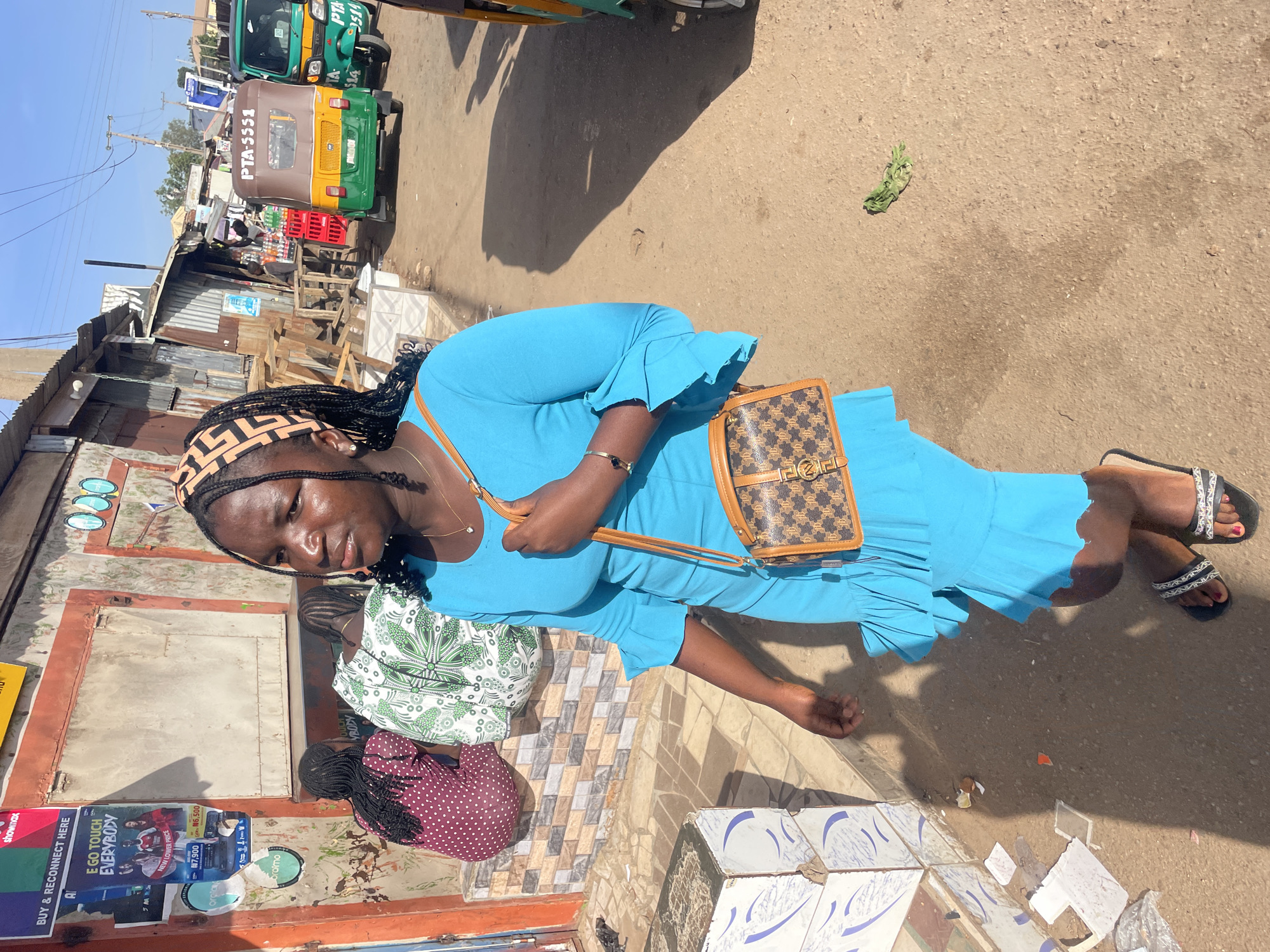 The image captures an outdoor street scene in Gura Top, Nigeria, under bright daylight with strong shadows indicating clear weather. The primary subject is a woman standing in the foreground, looking towards the viewer with a neutral expression. She wears a vibrant blue long-sleeved dress, black and white patterned slide sandals, and carries a small brown and black patterned handbag with a distinctive gold emblem. Her hair is styled in dark braids, partially covered by a patterned headscarf, and she wears a bracelet.
Behind her, two other women are partially visible, appearing to be sitting or crouching. One wears a green and white patterned top, and the other a maroon polka-dot top. They are positioned near rudimentary stalls or shops constructed from corrugated metal and wood.
The background features two auto-rickshaws (tuk-tuks) parked on a dirt road. One is brown and yellow with the text "PTA-5551" on its rear, while another green and yellow tuk-tuk displays "PTA 514". Red crates are stacked near the stalls.
Posters and advertisements are visible on a wall or structure to the left, with text including "SHOWMAX", "BUY & RECONNECT HERE", "E GO TOUCH EVERYBODY", "NEW LOWER PRICE", "₦7,900", "₦6,500", and "oraimo". The ground comprises dirt, broken tiles, and some paved sections, depicting a bustling, informal market or commercial area.