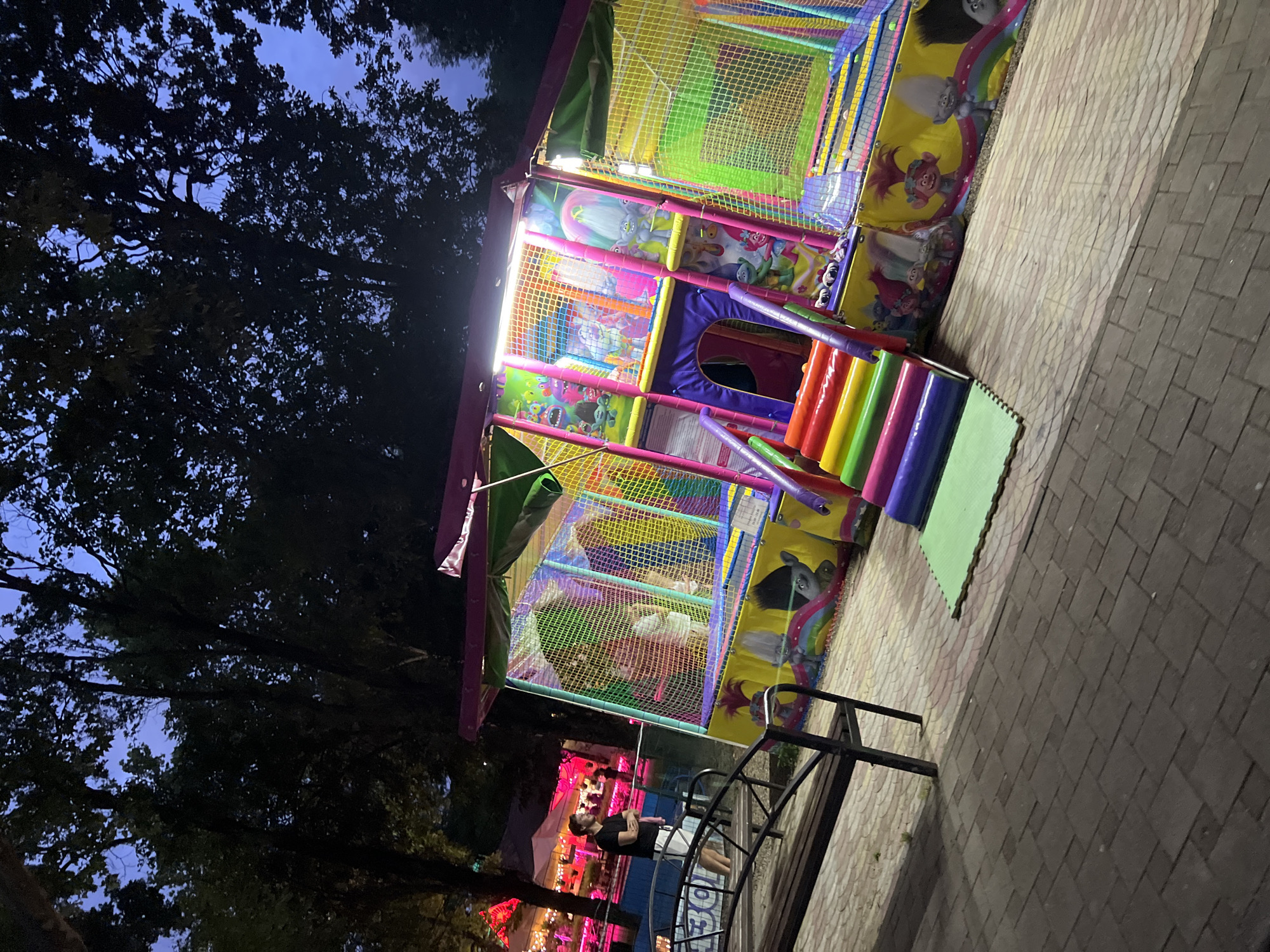 The image depicts an outdoor children's playground attraction with a Trolls theme, illuminated by artificial lights, suggesting it is evening or nighttime. The attraction features colorful netting, slides, and depictions of the Trolls characters. In the foreground, a man in a black t-shirt and white shorts stands near a park bench. The ground is paved with interlocking stones. In the background, trees with dark foliage and a slightly overcast sky are visible, along with distant illuminated structures that may indicate a nearby entertainment venue or park. The overall scene suggests a public recreational area. While the image does not explicitly state its location, it is described as being in Krasnodar, Russia.