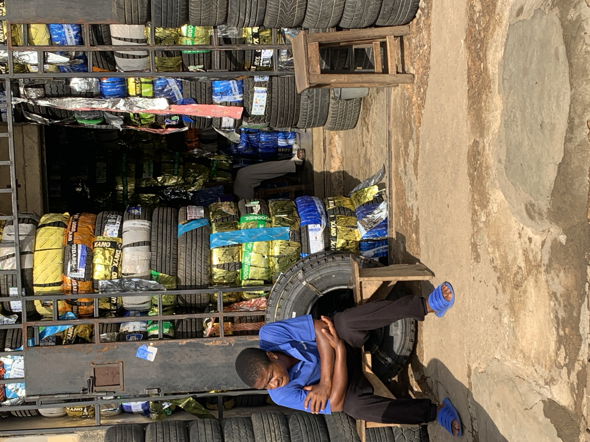 A man in a blue shirt and dark pants is sitting on a wooden stool in front of a tire shop in New Stadium, Nigeria. The shop is filled with stacks of tires wrapped in colorful packaging, with some tires on display on shelves. The man appears to be resting or waiting, with his arms crossed. The ground is concrete and shows signs of wear and age.