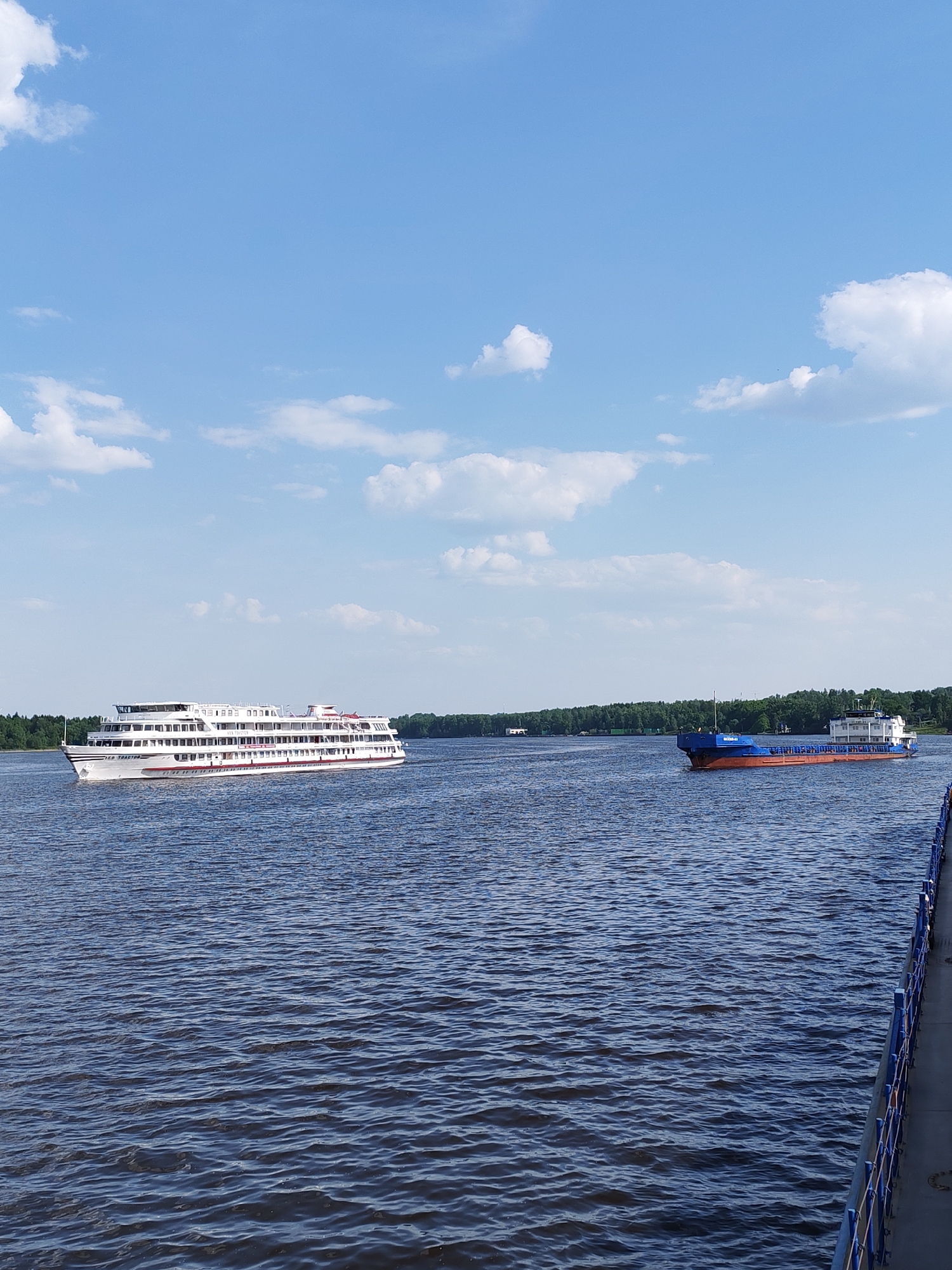 The scene depicts a body of water, likely a river, under a partly cloudy sky. Two vessels are visible on the water. On the left is a large, white passenger ship with multiple decks. On the right is a smaller cargo ship, predominantly blue with some red on the hull. In the distance, trees line the shore on both sides of the river. A portion of a blue and gray structure, possibly a pier or railing, is visible in the lower right corner of the frame. Based on the city of Ermolino, Russia, it is most likely the Protva River. There is no evidence of people in the image.