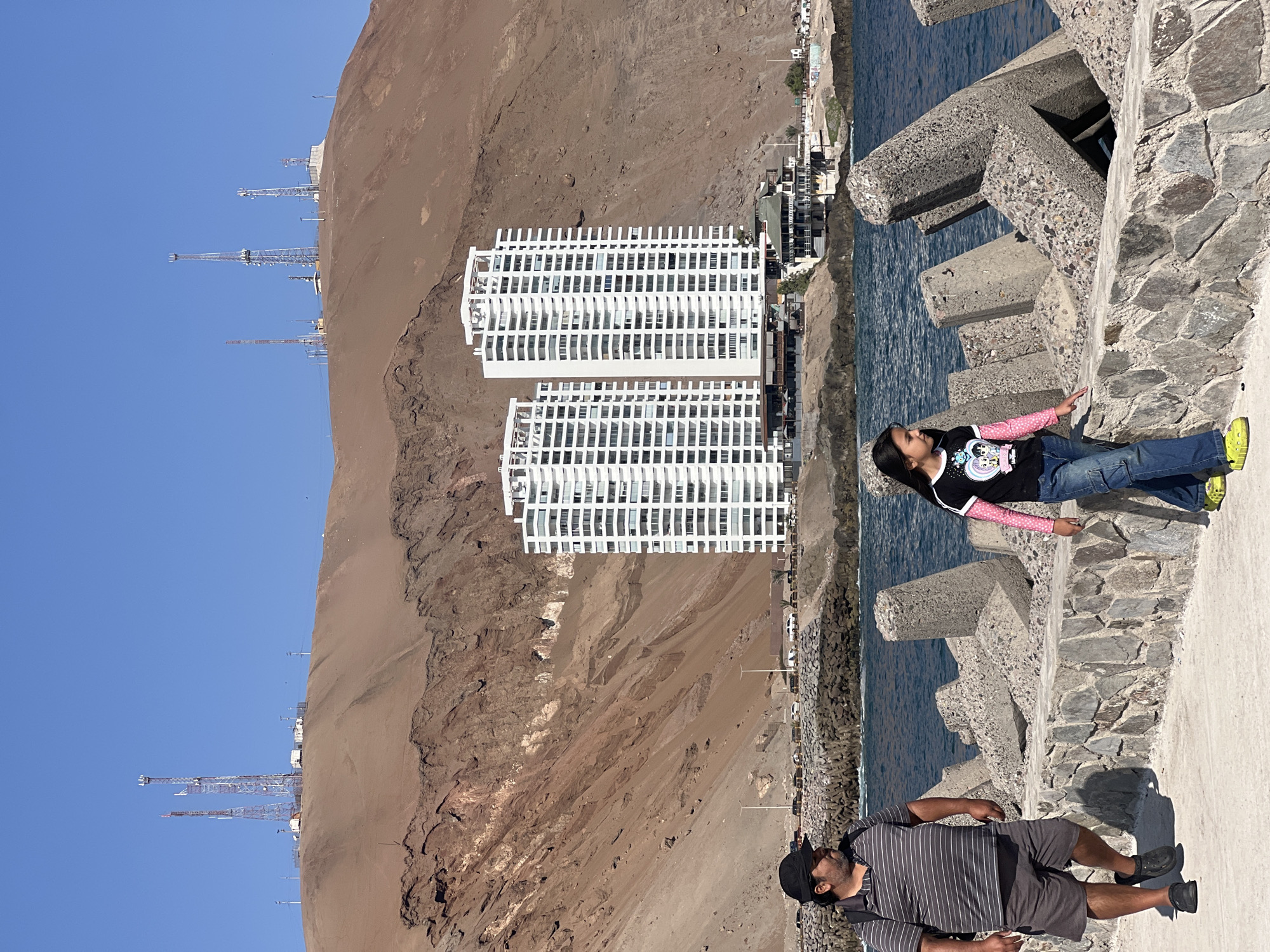 A scene in Arica, Chile, shows a landscape where the desert meets the Pacific Ocean. Two white high-rise buildings stand near the edge of a reddish-brown, eroded hill, with towers visible further up the hill. The sea's edge is lined with a concrete structure designed to break waves.
A man wearing a striped shirt, shorts, and a black cap lies on the concrete structure. Beside him, a child with long hair lies on the same structure, wearing a black shirt with a colorful print, jeans, and bright green shoes. Both are positioned near the water's edge, with one appearing to be looking towards the water. The photo is taken in bright daylight.