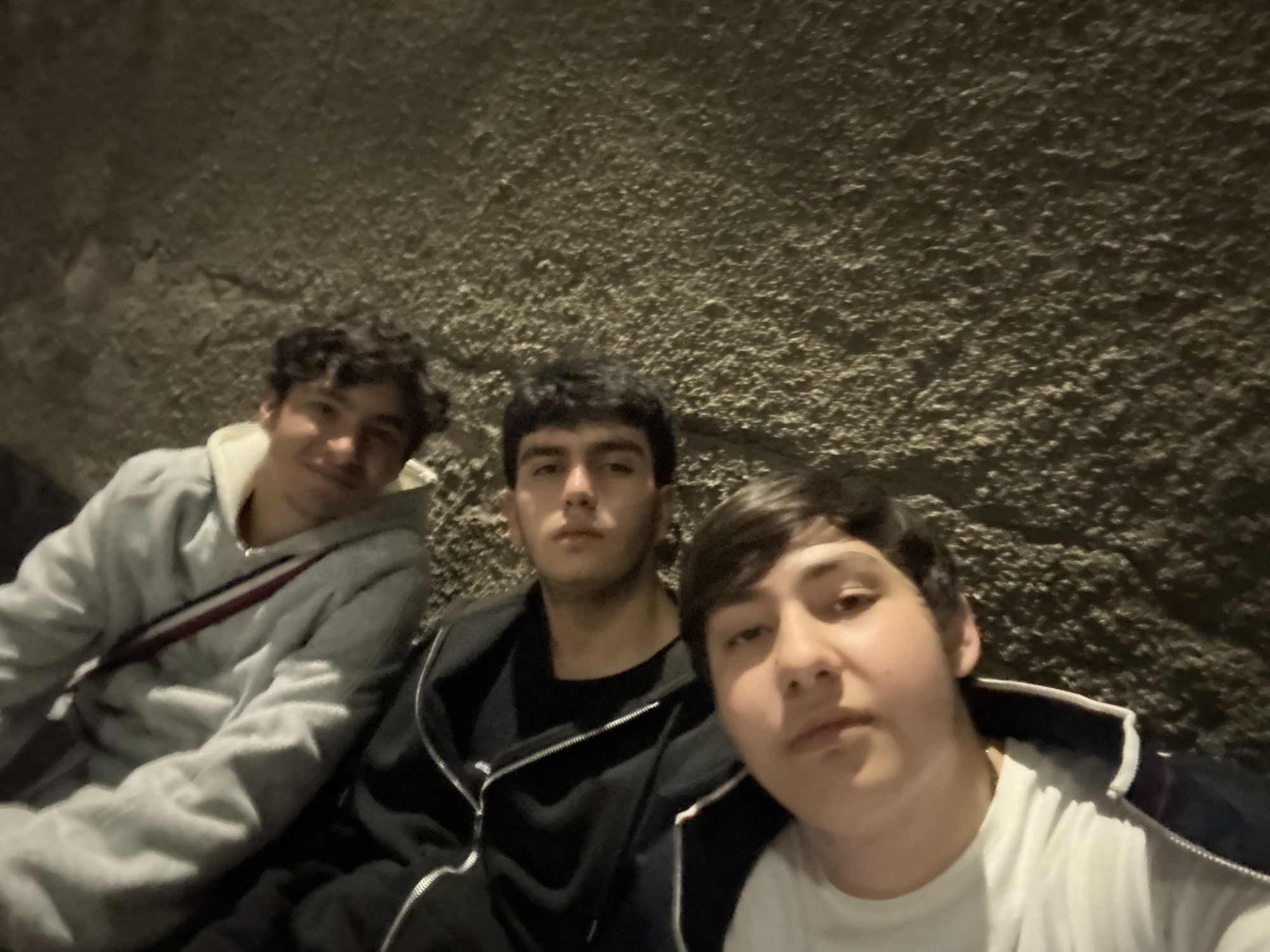 Three young males are posing for a selfie. The background is a rough, dark surface, possibly a wall. The male on the left is wearing a light gray hoodie with white and red details on the shoulder. The male in the center is wearing a black hooded sweatshirt over a black t-shirt. The male on the right is wearing a white t-shirt. The setting is dimly lit, suggesting an indoor or nighttime environment. Due to the limited context, the location in Yerevan, Armenia cannot be determined.