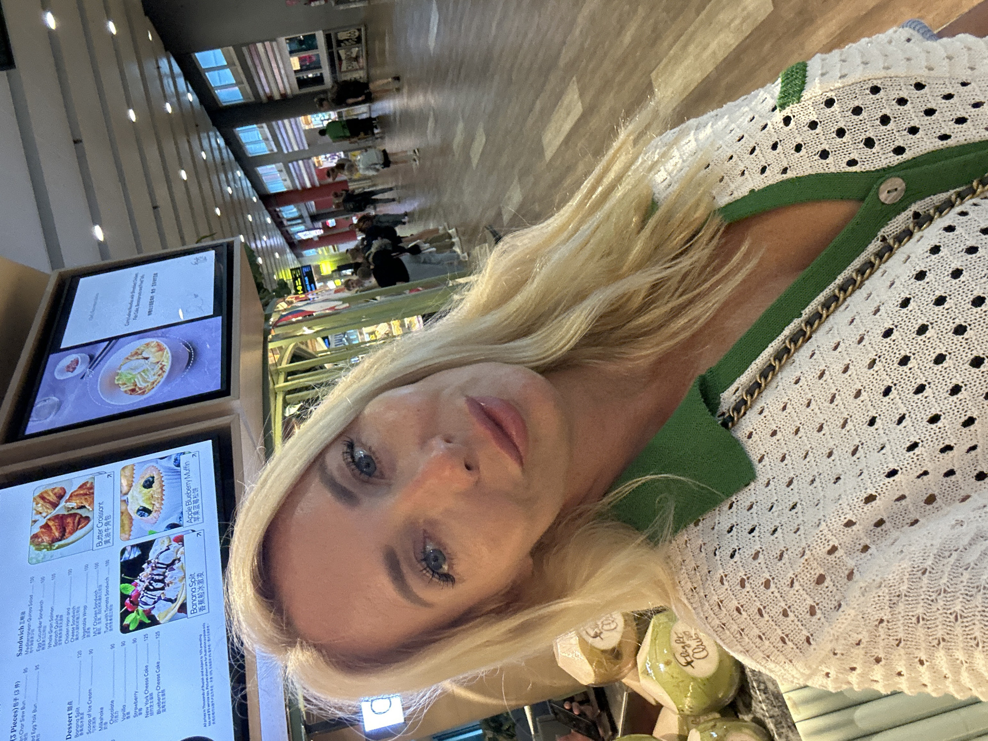 A blonde woman is taking a selfie in an indoor setting. She is wearing a white, open-knit sweater with green trim and a green collar, and a gold chain purse strap is visible across her chest. In the background, a digital menu board displays food items. To the left of the woman, several coconuts are stacked. The background shows a brightly lit walkway with other people walking in the distance. The overall environment appears to be a food court or a similar public indoor space, possibly a mall or airport terminal in Kuta, Indonesia.