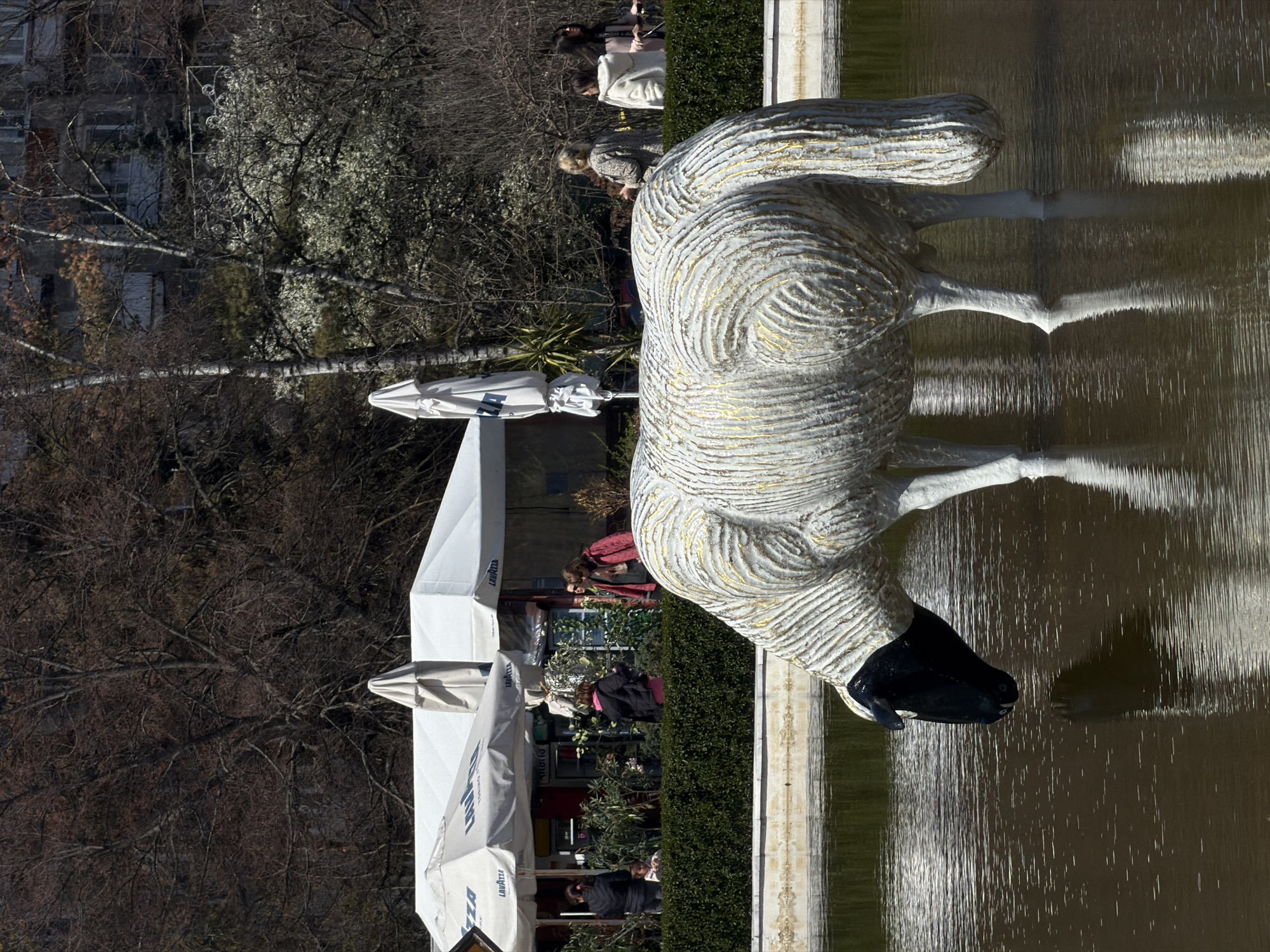 A statue of a sheep with a black head and white body with gold markings stands in a shallow pool of water. The statue's reflection can be seen in the water. Behind the pool, a row of people walk along a path. There are umbrellas and outdoor seating, possibly for a cafe or restaurant. Trees without leaves are on the left, and buildings are visible in the background. This is likely the sheep statue in the City Garden park in Sofia, Bulgaria.
