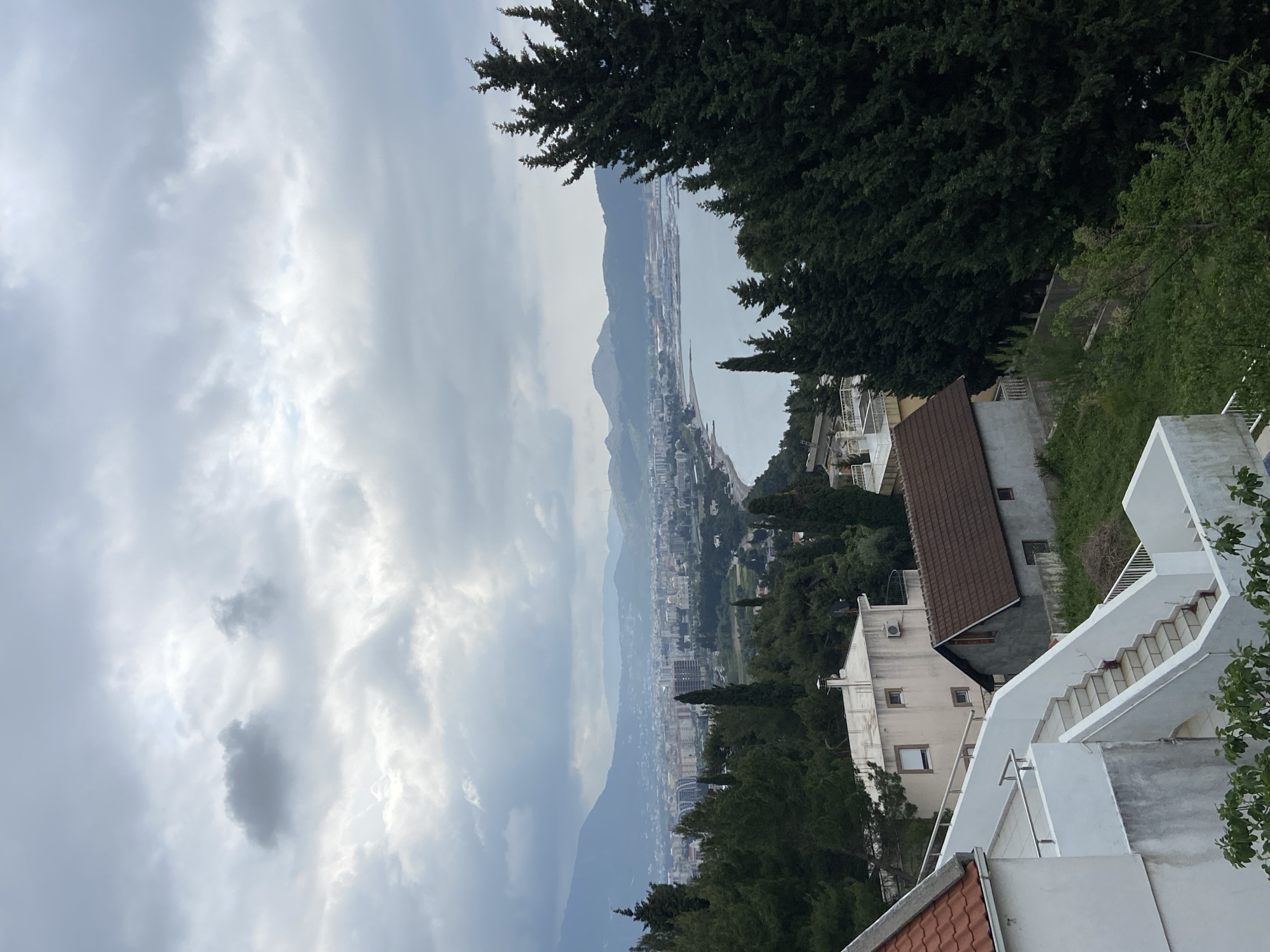 The image presents a panoramic landscape view of a coastal town or city, likely Šušanj, Montenegro, nestled between mountains and the sea, captured from an elevated perspective on a cloudy day.
The sky, occupying the upper portion of the frame, is heavily overcast with various shades of grey and white clouds, suggesting diffused daylight. In the distant background, a range of mountains or large hills appears hazy and partially obscured by the atmospheric conditions, rising behind the coastal area.
The midground reveals the expansive urban development stretching along the coastline. Numerous buildings of varying sizes and colors line the sea, with some areas appearing more densely built up. The sea, calm and grey under the cloudy sky, extends into the distance, with a long stretch that appears to be a beach or promenade visible along its edge. Further down the coast, what might be a port or industrial area with structures and cranes can be discerned.
In the foreground, closer to the viewer, are elements of the hillside from which the photo was taken. Several residential buildings with white facades and reddish-brown or dark brown tiled roofs are visible. A distinctive feature is a multi-level white concrete staircase with metal railings, descending sharply. Lush green vegetation, including tall, dark conifer trees (likely cypress trees, characteristic of the Mediterranean) and other leafy trees, densely covers the slopes, framing the urban and natural landscape.
The weather is distinctly cloudy and overcast, contributing to the soft, diffused lighting across the scene. The time of day appears to be daytime, but not under direct sunlight. No specific human activity is visible, and there is no discernible text within the image. The overall impression is one of a serene yet dramatically clouded coastal landscape.