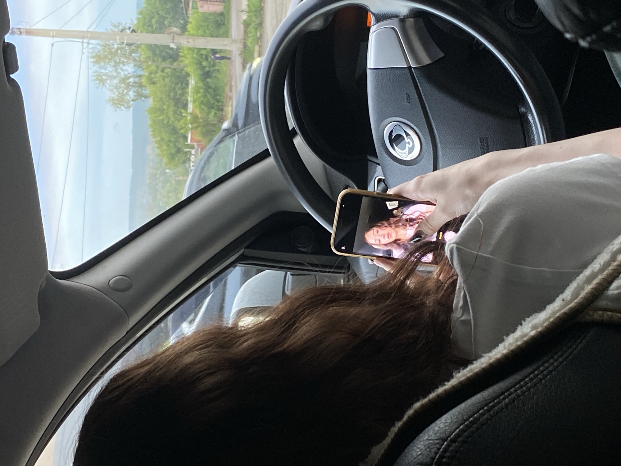 The image shows the interior of a car, looking out the passenger window. A person's hand is holding a smartphone, displaying a video call with a young woman. The woman in the video call has long, wavy brown hair and is wearing a pink top. The person holding the phone also has long, wavy brown hair.
Outside the car window, there is a view of a road lined with trees and some buildings. The sky is bright, suggesting it is daytime, possibly late morning or afternoon. The overall scene appears to be casual and everyday, with the focus on the interaction occurring on the phone. There is no visible text in the image. The context provided states the location is Votkinsk, Russia.