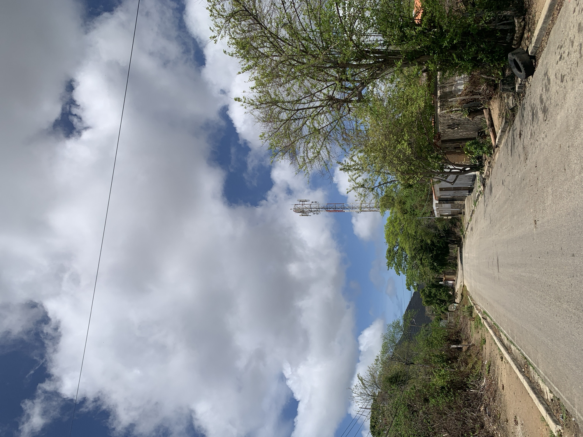 This is a ground-level, eye-level shot of an empty, paved road leading into the distance. The road is flanked by sparse vegetation and what appear to be small, rustic buildings on the right side. A cellular tower is visible in the background, to the left of a large tree. The sky is partly cloudy, with large white cumulus clouds against a blue backdrop. The overall impression is one of a rural or semi-urban setting, likely in a warm climate. The scene appears to be located in the city of Santa Ana, Venezuela.