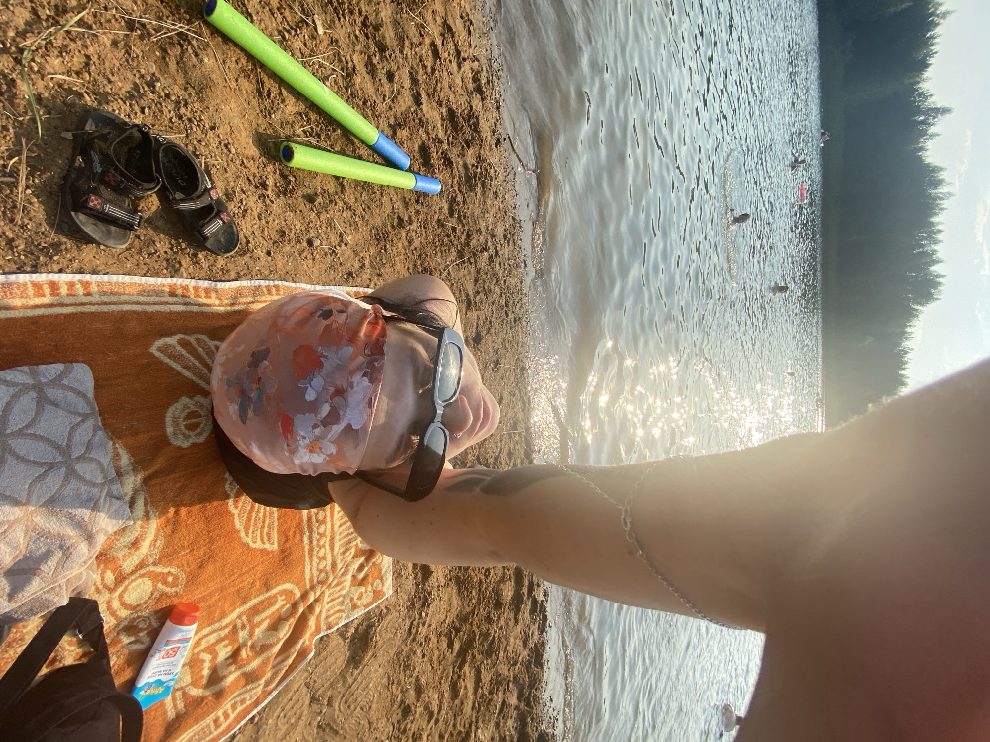 This is a selfie taken at a beach in Novyi, Russia. The image shows a person lying on a sandy beach, with a lake and forest in the background. The person is wearing sunglasses and a floral-patterned face mask. Two green and blue pool noodles are lying on the sand next to the person, along with a pair of black sandals. An orange towel with a patterned design is spread out on the sand. A tube of sunscreen is visible on the towel. The lake is calm with a few people swimming in the distance. The sun is shining brightly, creating reflections on the water. The overall scene suggests a relaxed summer day at a lake.