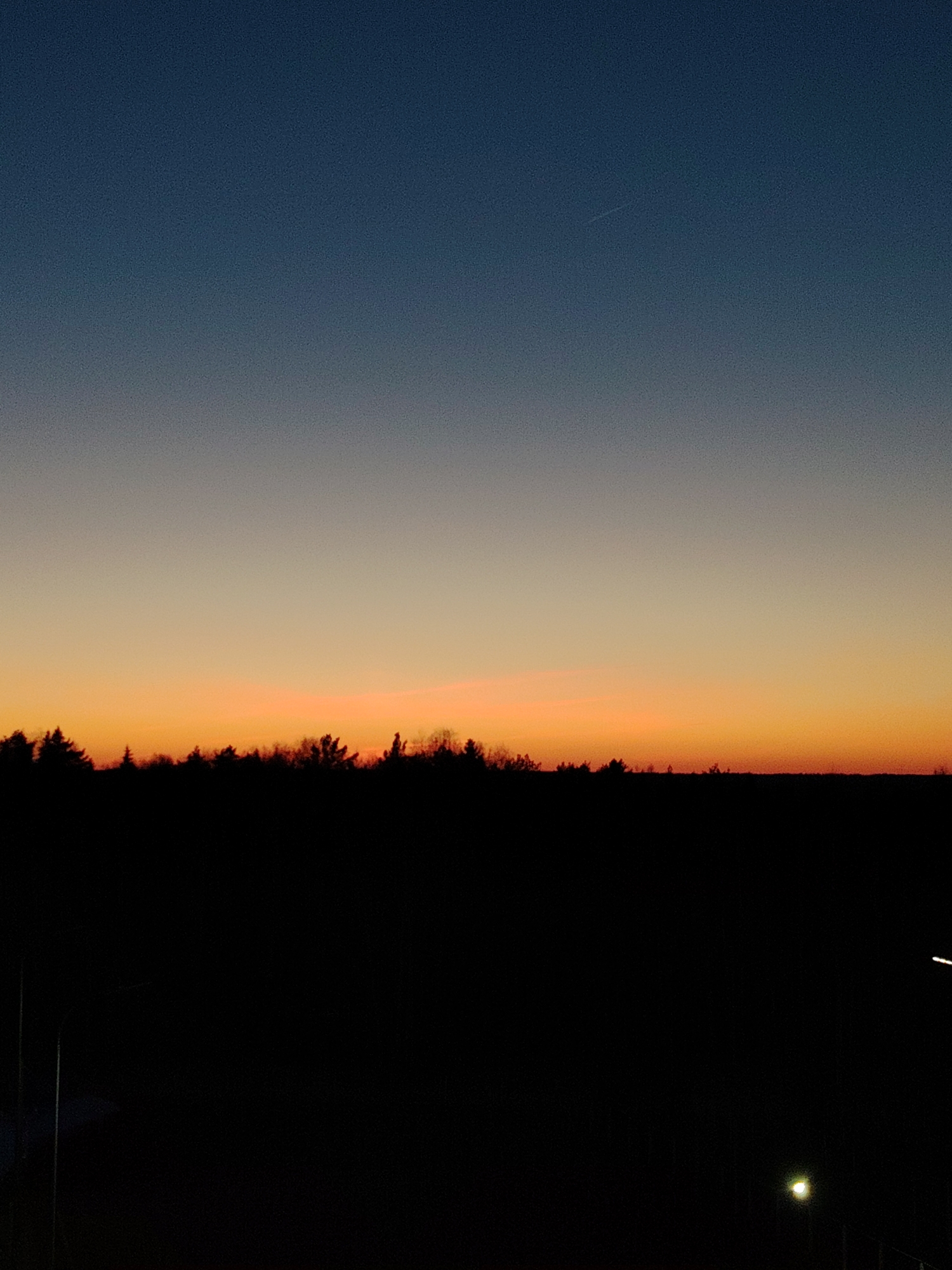 The image captures a twilight scene in Марковский, Russia, characterized by a gradient sky transitioning from dark blue at the top to orange near the horizon. A silhouette of trees and possibly other vegetation is visible against the bright horizon. The foreground is largely dark, suggesting a low-lit area. A few streetlights are visible in the lower right of the frame.