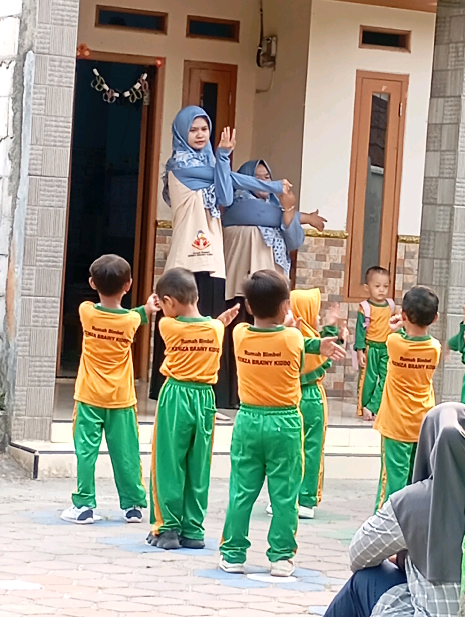 In Babelan, Indonesia, in front of a building with beige walls and brown doors, a group of children in matching orange shirts and green pants are gathered, seemingly engaged in a physical activity guided by two adult women. One woman, wearing a blue hijab and a light-colored smock over her clothing, stands on the left, demonstrating movements with her arms raised. The other woman, also in a hijab, stands to her right, her arms extended. The children are facing the women, with their arms outstretched in various poses, suggesting they are following instructions. The ground is paved with interlocking bricks. Some children are wearing backpacks. The overall scene appears to be an outdoor class or activity session for young students.