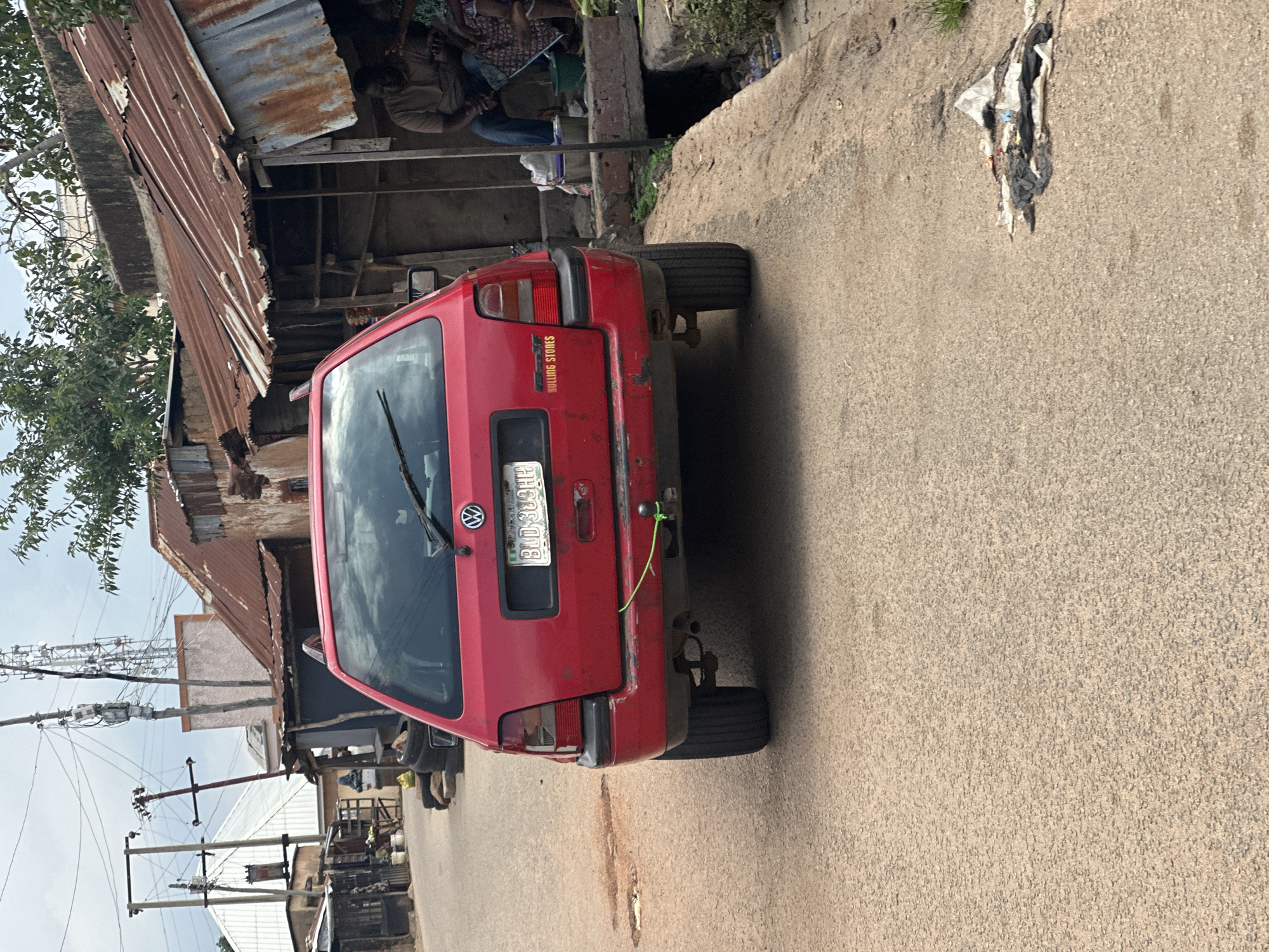 An eye-level, medium shot captures the rear of a red Volkswagen Golf parked on a paved street in what appears to be a rural or less developed area. The car's license plate reads "BLD-389HH." A green rope is tied to the trailer hitch on the rear bumper.
To the right of the car, a narrow drainage ditch runs along the side of the road. On the far side of the ditch, a man in a light brown shirt and blue jeans is seated next to another person. Behind them, a weathered structure with a corrugated metal roof is visible. In the background, more buildings with similar roofing materials line the street, interspersed with utility poles and communication towers. The sky above is overcast. Scattered debris is present on the ground to the right of the car.