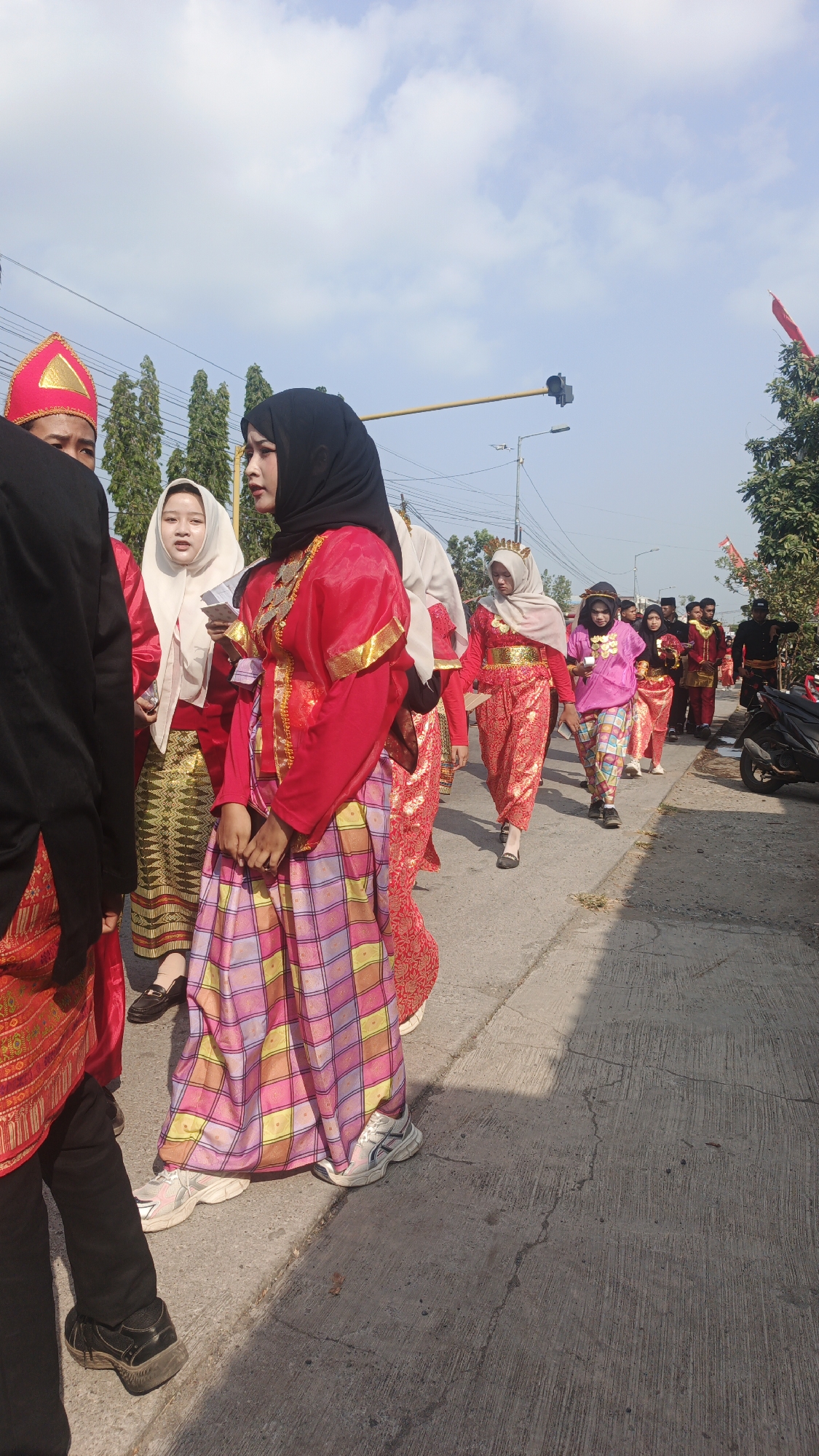 In Sumengko, Indonesia, a group of people are walking in a procession along a paved road under a bright sky with scattered clouds. The individuals are dressed in traditional attire, predominantly red and gold, with some wearing headscarves. The procession appears to be moving from left to right in the frame. In the background, there are trees and utility poles. To the right of the road, a motorcycle is parked. The scene is captured during daylight hours.