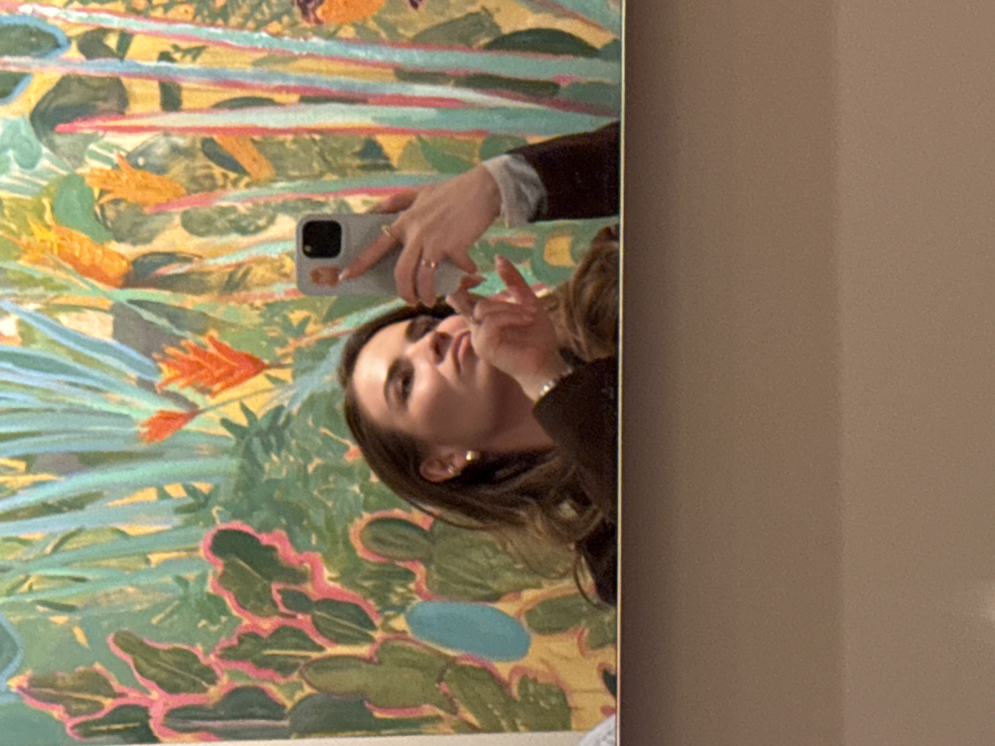 A woman takes a mirror selfie with a phone in front of a botanical painting. The painting is comprised of vivid colors like yellow, orange, green, pink, and blue. The woman is wearing a dark jacket and gold earrings. The location cannot be determined from the information provided.