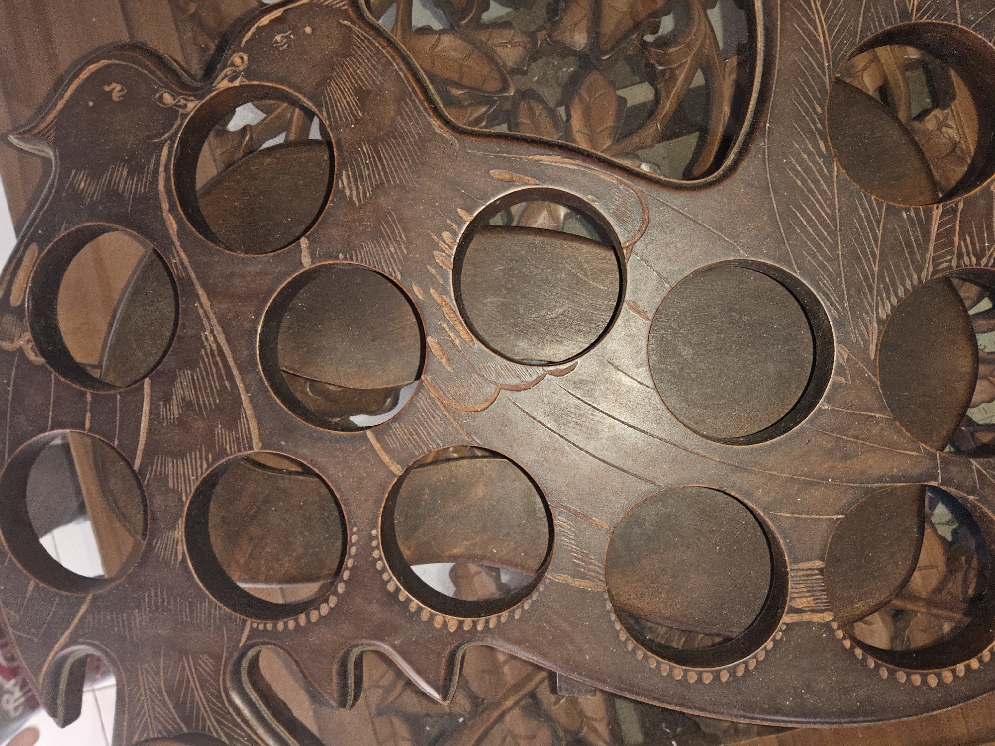 The image shows a close-up, top-down view of a dark brown wooden item with several circular cutouts. The wood is carved with intricate patterns, including what appear to be stylized leaves and lines resembling feathers. The circular openings reveal a blurred background, suggesting the item is part of a larger structure or display. The lighting is dim, casting shadows that highlight the texture and carvings of the wood. The item is likely a decorative piece or a functional holder. There is no visible indication of the location being in the city of Payagaboh, Indonesia, nor are there any people, actions, or interactions depicted in the image.