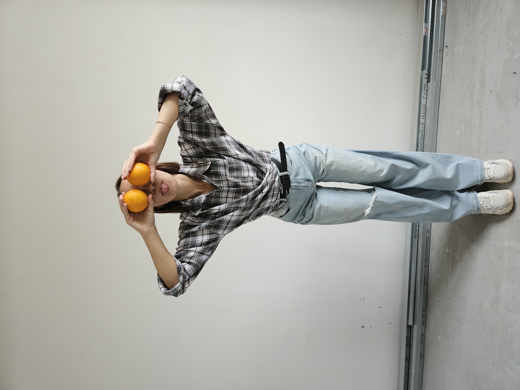 A woman lies on her side on a grey concrete floor against a white wall, holding an orange in each hand in front of her eyes. She is wearing light blue jeans with a rip in the left leg and a plaid button-up shirt. Metal studs lean against the wall beside her feet. The image is rotated counter-clockwise. Based on the OCR, the location is Chaikovsky, Russia.