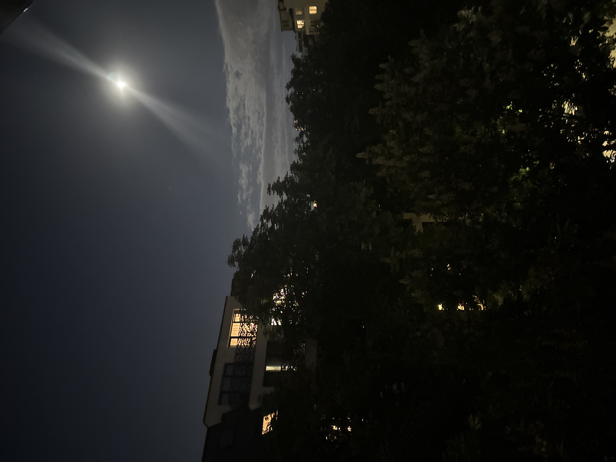 The image shows a night scene in Sofia, Bulgaria. The moon is prominent in the upper left, with light rays emanating from it. A cloud formation stretches vertically in the middle of the image. Two apartment buildings are visible, one on the lower left with lights on in several windows, and another in the upper right, also with illuminated windows. Trees are in the foreground and right, partially obscuring the buildings. The overall lighting is dark, with the moon and building lights providing the primary sources of illumination.