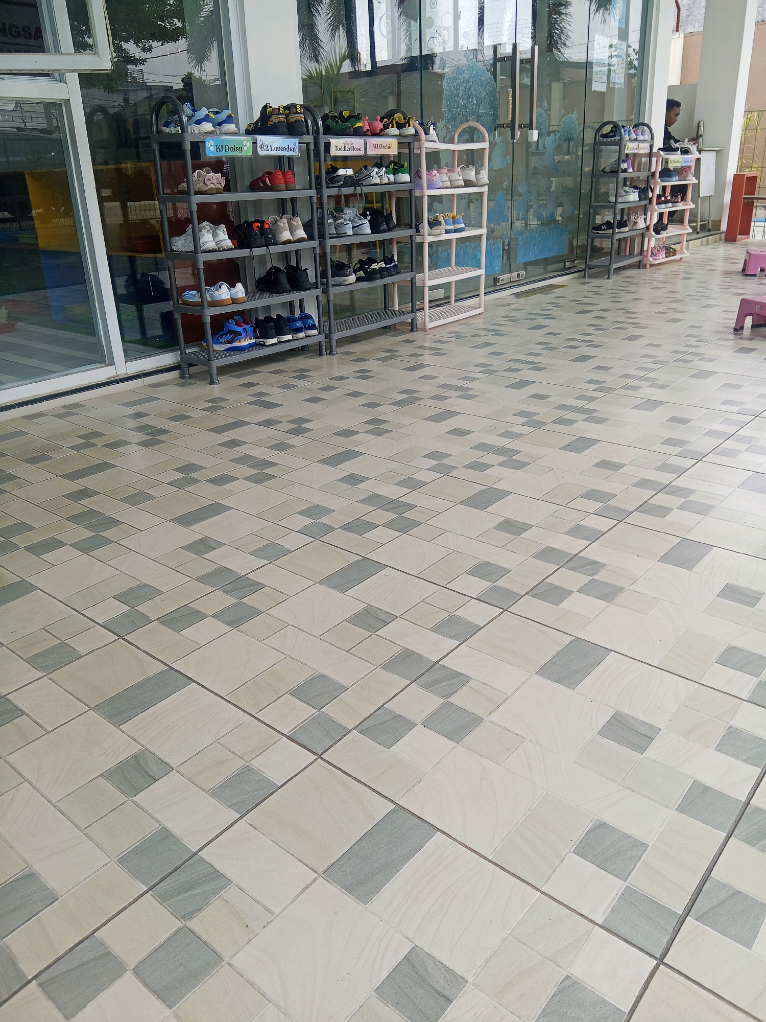 This image depicts an outdoor tiled walkway in front of a building with large glass doors. In the foreground, the tiled floor features a pattern of light beige and grey squares. Beyond the walkway, several multi-tiered shoe racks are visible, holding various pairs of children's shoes. The shoe racks are organized and appear to be for a school or daycare facility. The building's facade includes large windows and a doorway. In the background, a person can be seen standing near a shoe rack. The scene suggests a tidy and organized environment, likely associated with a children's educational institution. The location is identified as being in the city of Lubuklinggau, Indonesia, based on the provided context.