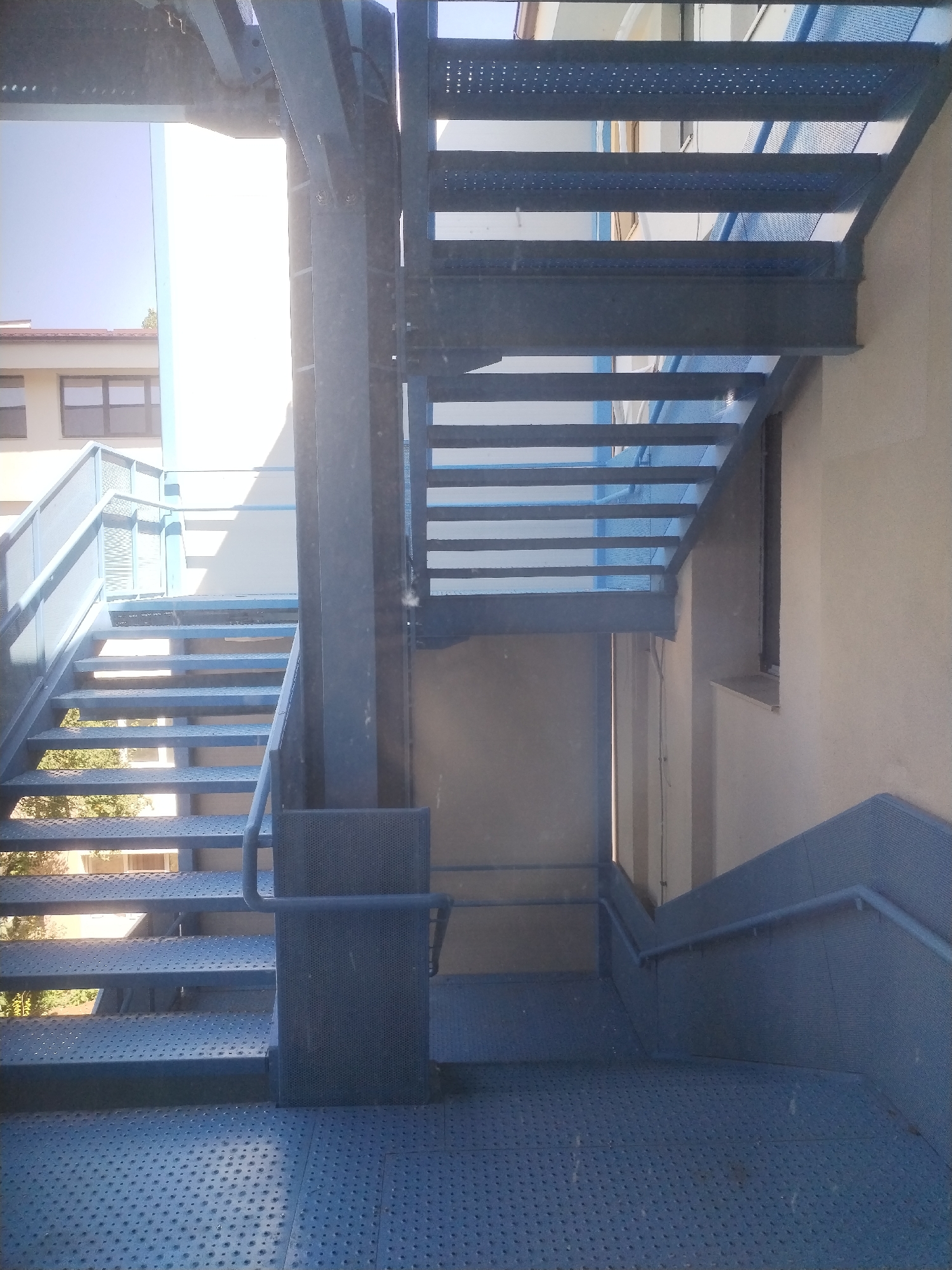 The image shows an outdoor metal staircase painted blue. The staircase has perforated metal treads and risers, with metal railings on the sides. The staircase appears to be part of a building's external fire escape or access route. In the background, a lighter-colored building with windows is visible, along with some trees. The overall scene is an exterior view of architectural elements, likely in an urban setting. There are no people or animals visible in the image, nor are there any specific landmarks or indications that would place this scene in Bucharest, Romania.