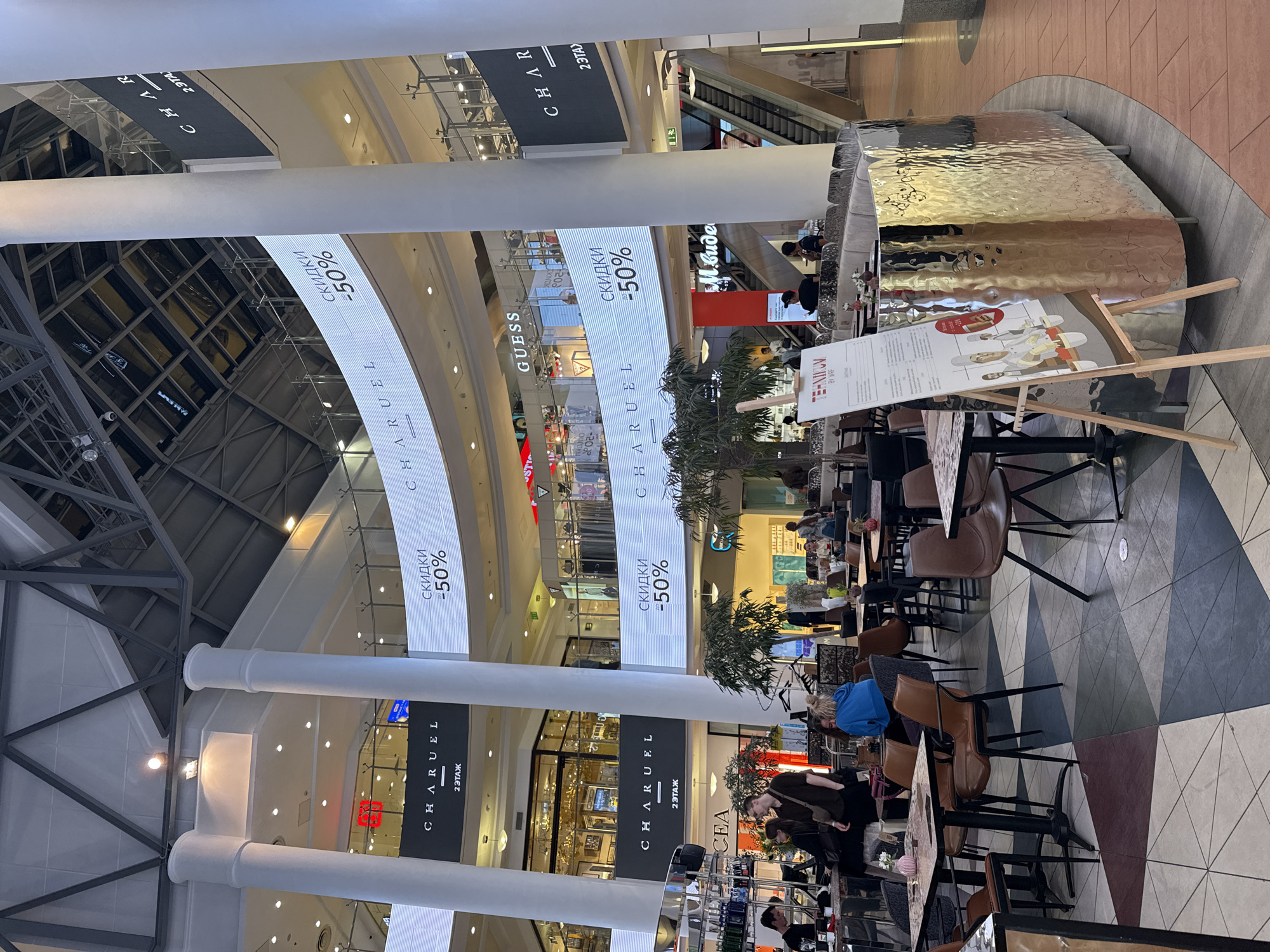 The image depicts the interior of a multi-level shopping mall, likely in Moscow, Russia, given the Cyrillic signage. The architecture features large white cylindrical columns supporting elevated walkways and a complex geometric ceiling structure with visible metal supports and lighting. Large, curved digital displays are prominent, showing advertisements with Cyrillic text, including "СКИДКИ -50%" (Discounts -50%) and "CHARUEL".
In the foreground, several tables and chairs are arranged in a cafe or restaurant seating area. People are seated at tables, some appearing to be dining or socializing. A group of people are standing near a counter on the left side of the image, suggesting a service area. A person in a blue top is seated at a table, and another person in a brown top is standing nearby.
A display easel with a menu or advertisement is positioned in the mid-ground, featuring illustrations of chefs and food items. The flooring is a patterned mosaic of geometric shapes in various neutral tones. In the background, escalators are visible, indicating movement between levels. Several shops and store facades are discernible on different levels, with brand names like "GUESS" and "CHARUEL" visible. The overall atmosphere is that of a busy, modern commercial space.