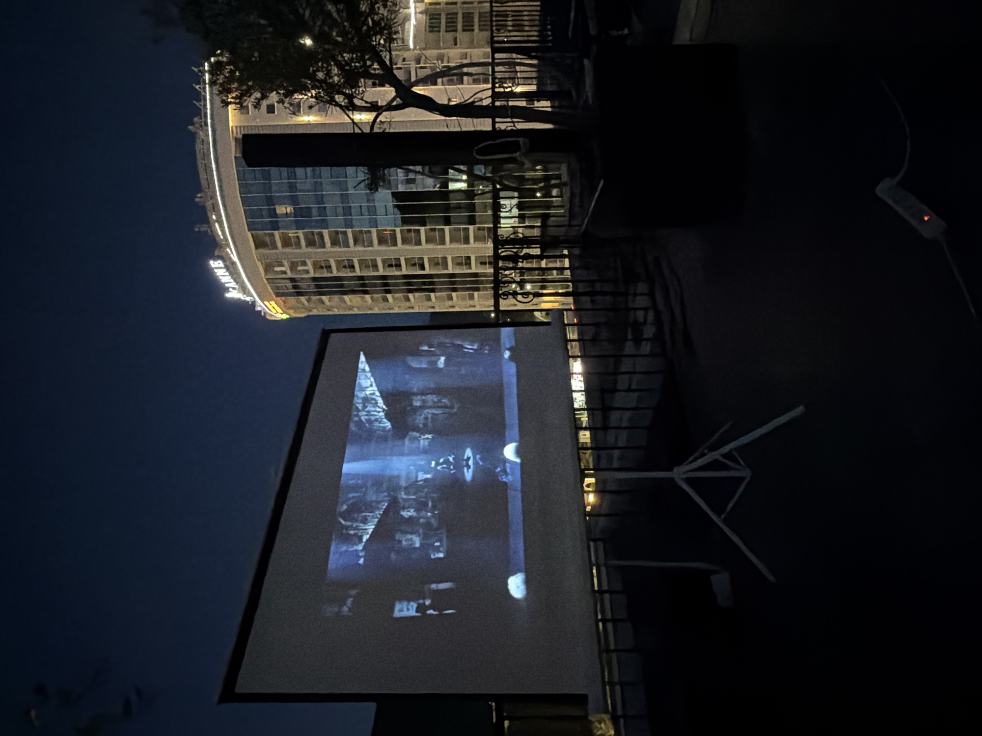 Here's a description of the image:
The image shows an outdoor movie setup at night. A large projector screen displays a film, reflecting the light onto its white surface. Behind the screen is a wrought-iron fence, beyond which is a tall building with a curved façade bearing the illuminated letters "CANNES". A tree branch stretches across the upper part of the frame, partially obscuring the building. To the right of the screen, a white electrical extension cord can be seen. The image is taken in the city of Budva, Montenegro.