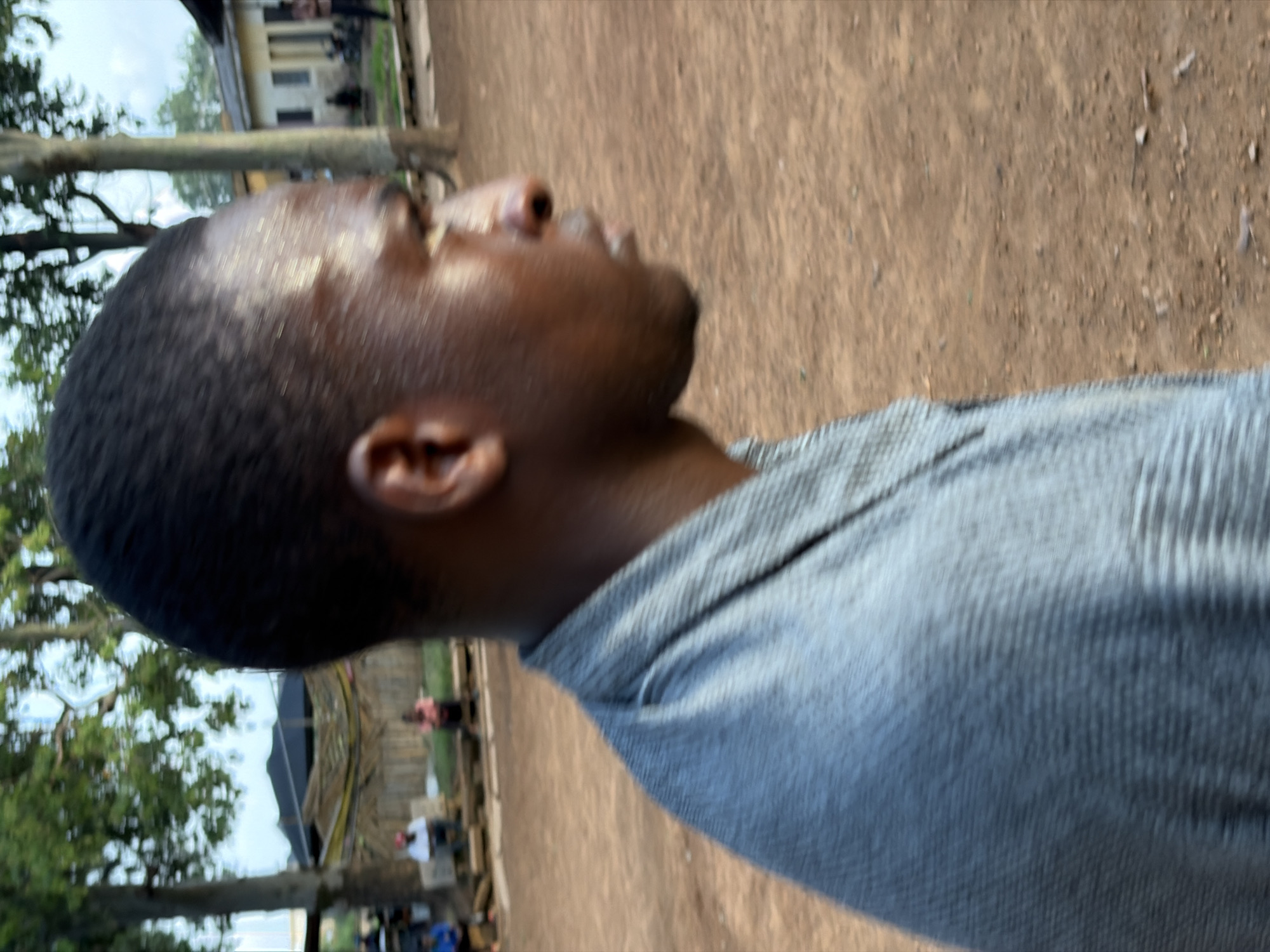The image shows a close-up, side-profile view of a Black man's head and shoulders, wearing a gray, textured shirt. He is outdoors, with a dirt ground and some vegetation in the background, including trees and what appears to be buildings. The lighting suggests it is daytime. There are other people and structures visible in the background, including a building with a dark roof. Based on the visual context and typical scenes from Rafin Par, Nigeria, the image likely depicts a common street or community area.