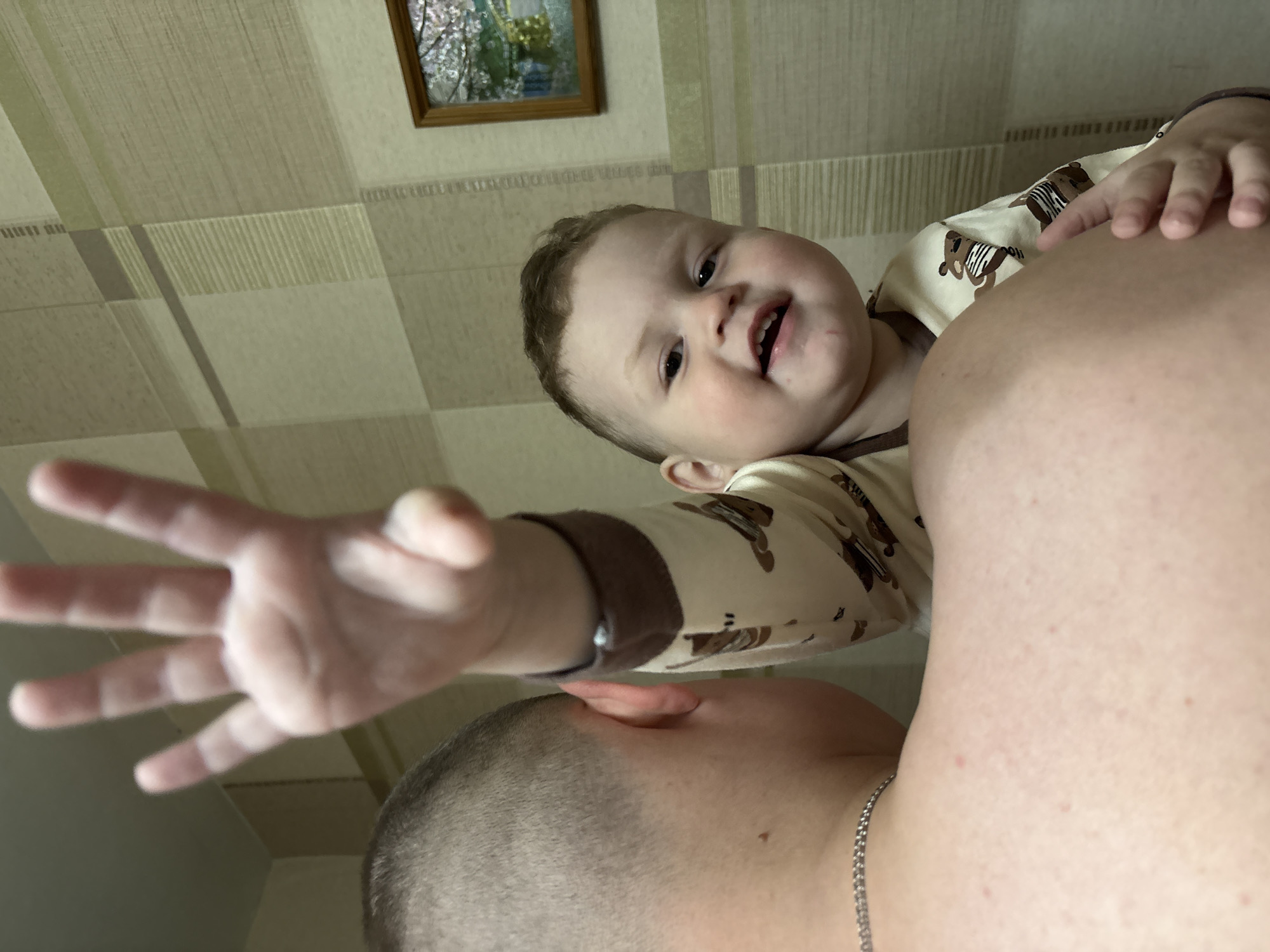 A man is holding a baby in his arms. The baby is smiling and looking at the camera. The man's hand is raised in the air, as if he is waving. The background is a wall with a picture frame. The location is unknown.