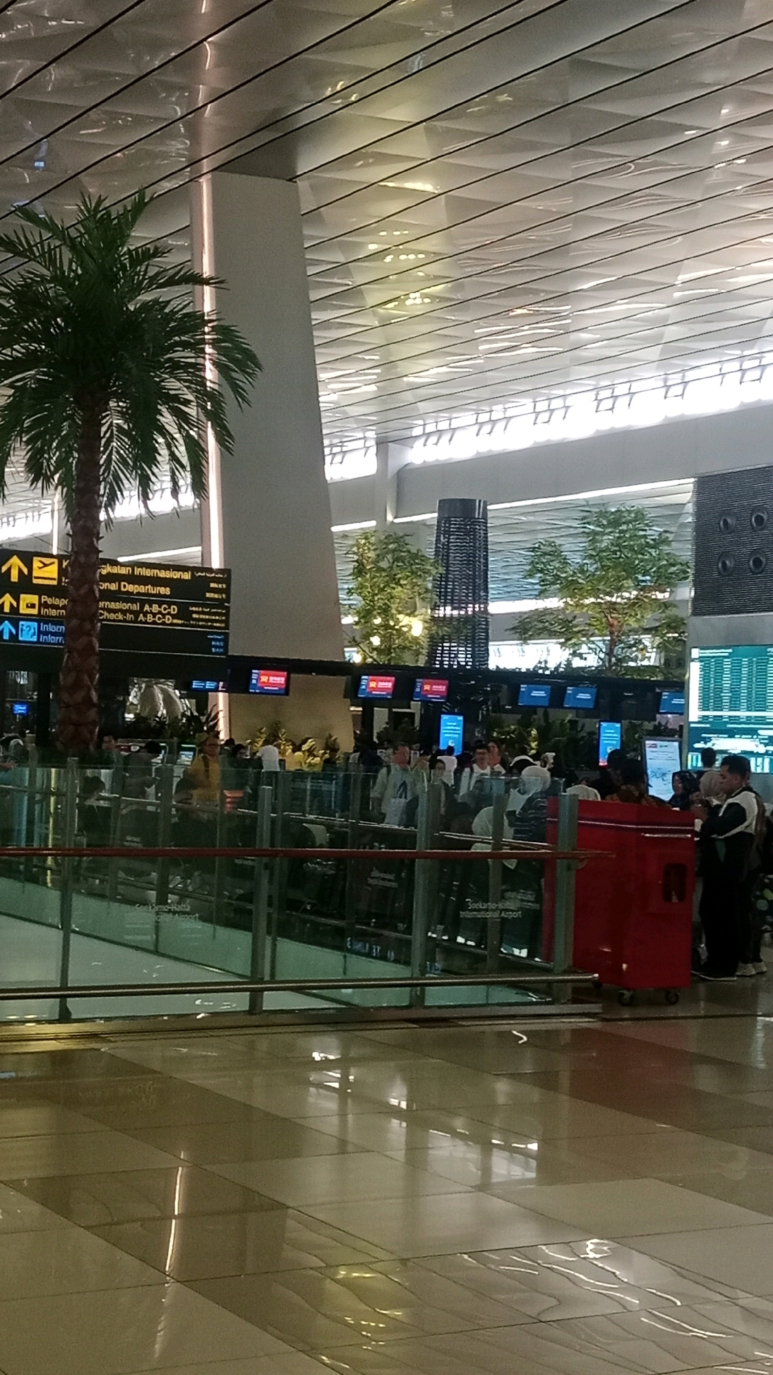 The image depicts the interior of an airport terminal. A prominent feature is a tall, slender column with a textured, dark cylindrical structure atop it, resembling a modern art installation or ventilation shaft. In the foreground, a glass barrier separates the viewer from a queue of people. The people are of various ages and ethnicities, dressed in casual attire, some are standing, others are in line. Behind the people, there are signs with flight information, and a large screen displaying data. A potted palm tree is visible to the left, and further back, artificial trees with small lights add to the ambiance. The ceiling is a complex geometric structure with integrated lighting. The floor is a polished surface, reflecting the ceiling lights and the surroundings. A bright red luggage cart is positioned on the right side of the image. The overall impression is one of a busy, modern international airport. The location is identified as Soekarno-Hatta International Airport, near Pajang, Indonesia.