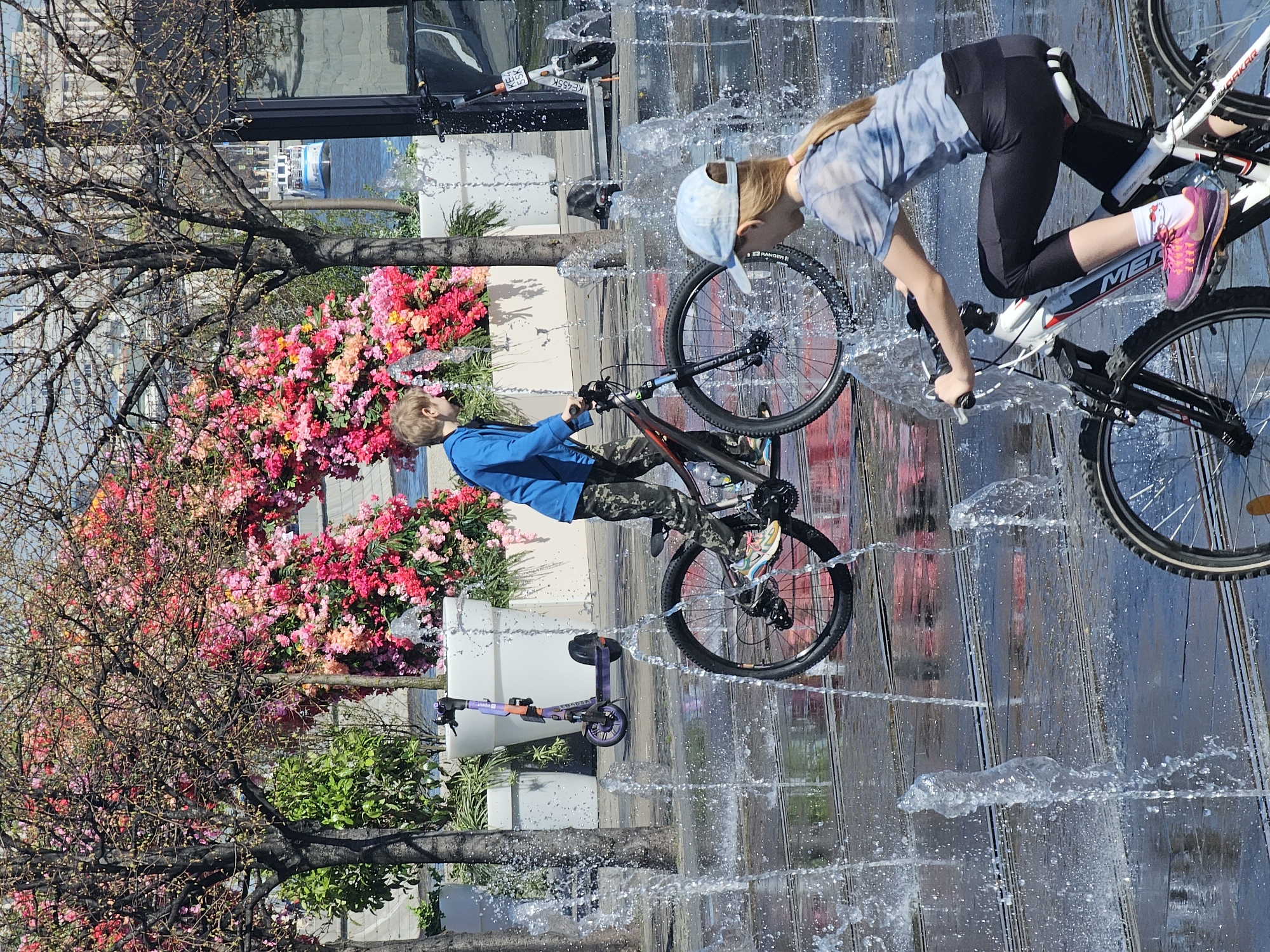 Two children ride their bikes in a water fountain in Moscow, Russia. The fountain is located in a plaza near flowering trees and decorative planters. A scooter is parked nearby. The girl is wearing a blue tie-dye shirt, black leggings, pink sneakers, and a blue cap. The boy is wearing a blue jacket and camouflage pants.